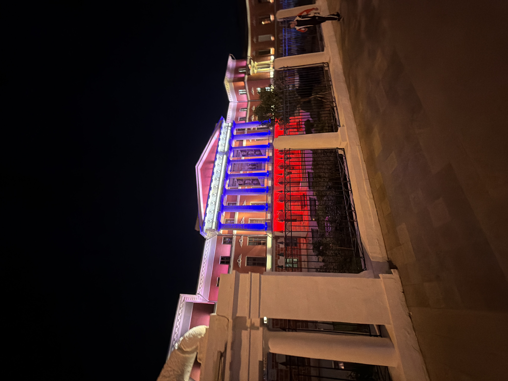 Here is a detailed description of the image:
The image captures a building in Moscow, Russia, illuminated at night with red, white, and blue lights that reference the Russian flag. The building is made of pinkish stone and features white columns. The front of the building is lit up with blue and red lights that alternate along its facade. A black, wrought iron fence is visible in front of the building. A person is standing on the sidewalk above, partially visible. The sky is dark, indicating it is night time. The image is slightly tilted. The building appears to be a notable public or governmental structure.
