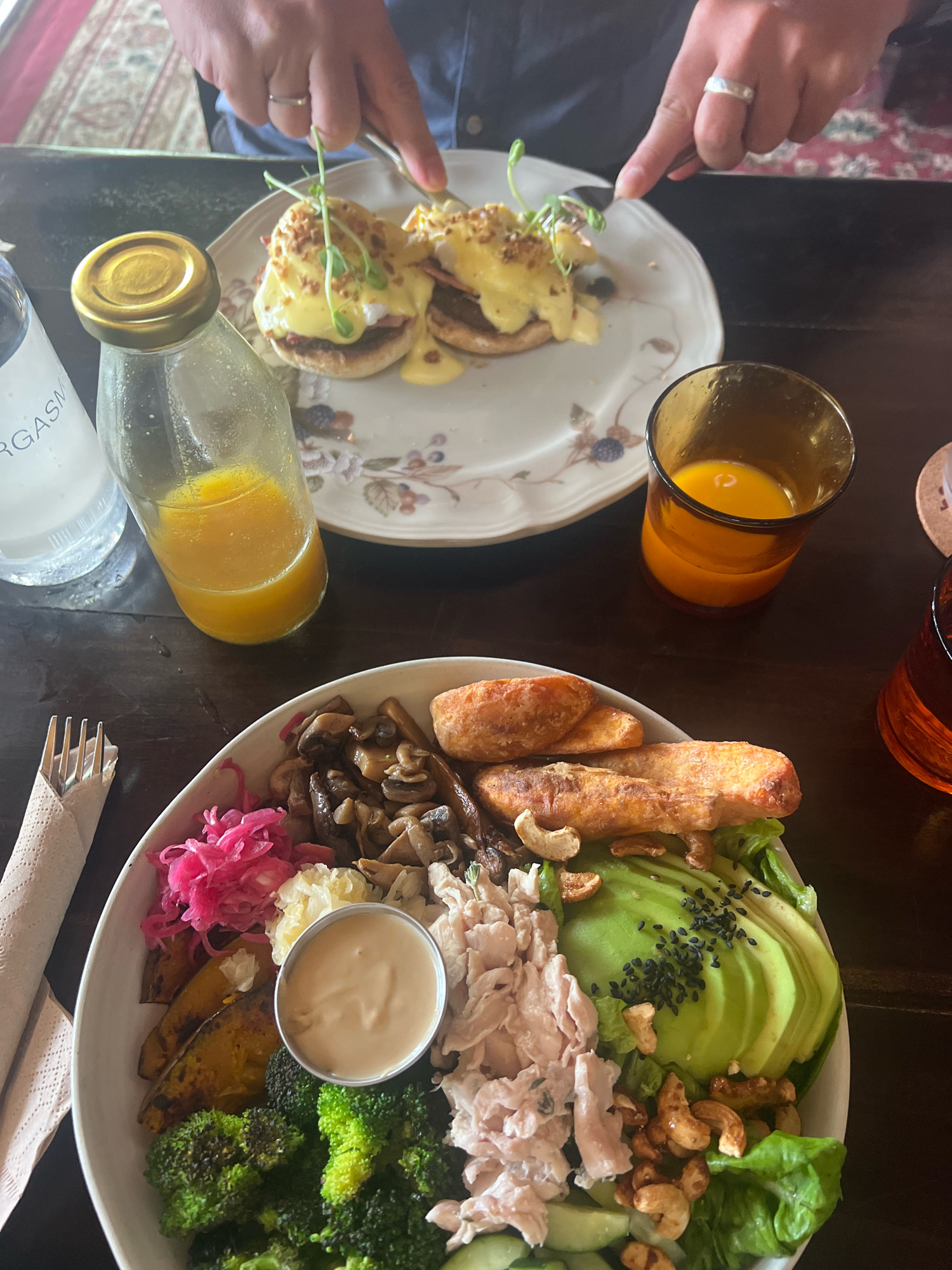 A table is set with two meals, one on a decorative plate and another in a large white bowl. In the foreground, the white bowl contains a salad with avocado, shredded chicken, cucumber, lettuce, roasted pumpkin, broccoli, mushrooms, and pickled red cabbage, with a small dish of dressing in the center. Cashews are scattered throughout the salad. To the left of the bowl, a fork is wrapped in a beige napkin. In the background, a decorative plate holds two English muffins topped with poached eggs and hollandaise sauce, garnished with microgreens. A glass bottle filled with orange juice and a glass of orange juice are also on the table. A person's hands are visible in the upper portion of the image, with one hand holding a fork and knife, preparing to cut into the meal on the decorative plate. The environment appears to be a dimly lit restaurant in North Kuta, Indonesia, with a wooden table and a patterned rug visible in the background.