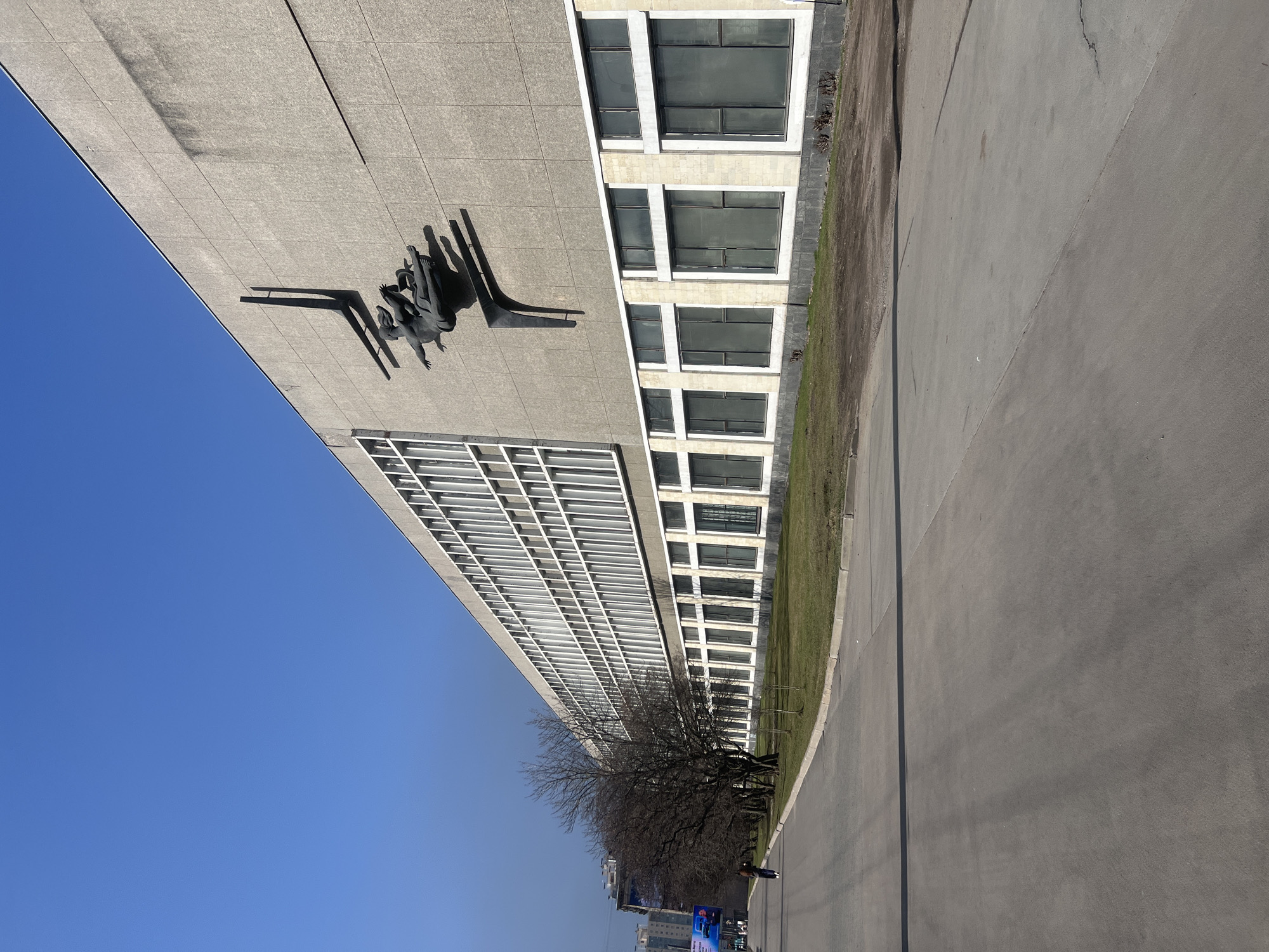The image presents an exterior view of the new building of the Russian National Library, located at Moskovsky Prospekt 165 in Saint Petersburg, Russia. The scene is captured on a clear, sunny day with a bright blue, cloudless sky.
The prominent structure on the right is a long, multi-story building with a light grey, paneled concrete facade. The upper section features a repeating pattern of numerous narrow, vertically oriented windows with white frames. The lower section displays larger, horizontally rectangular windows with dark glass, framed by white borders and separated by lighter-toned vertical sections.
Centered on the upper facade of the building is a large, dark-colored relief sculpture, known as "The Reader" (or "Knowledge") by Valentin Sveshnikov. It depicts two stylized human figures in dynamic poses, integrated with angular, abstract forms that project from the wall. The sculpture casts a long, sharp shadow diagonally upwards to its left, indicating strong sunlight from the right.
In the foreground, a wide, grey asphalt roadway stretches parallel to the building. To the left of the road, a narrow strip of unkempt grass and bare-branched deciduous trees runs alongside the building. One individual, dressed in dark attire, is visible walking away from the camera on the paved surface to the far left, near the line of trees. In the distant background on the left, urban infrastructure elements, including a large electronic billboard displaying a blue advertisement and parts of other buildings, are partially visible. The overall environment suggests an urban setting during daylight, likely in early spring due to the bare trees and patchy grass.