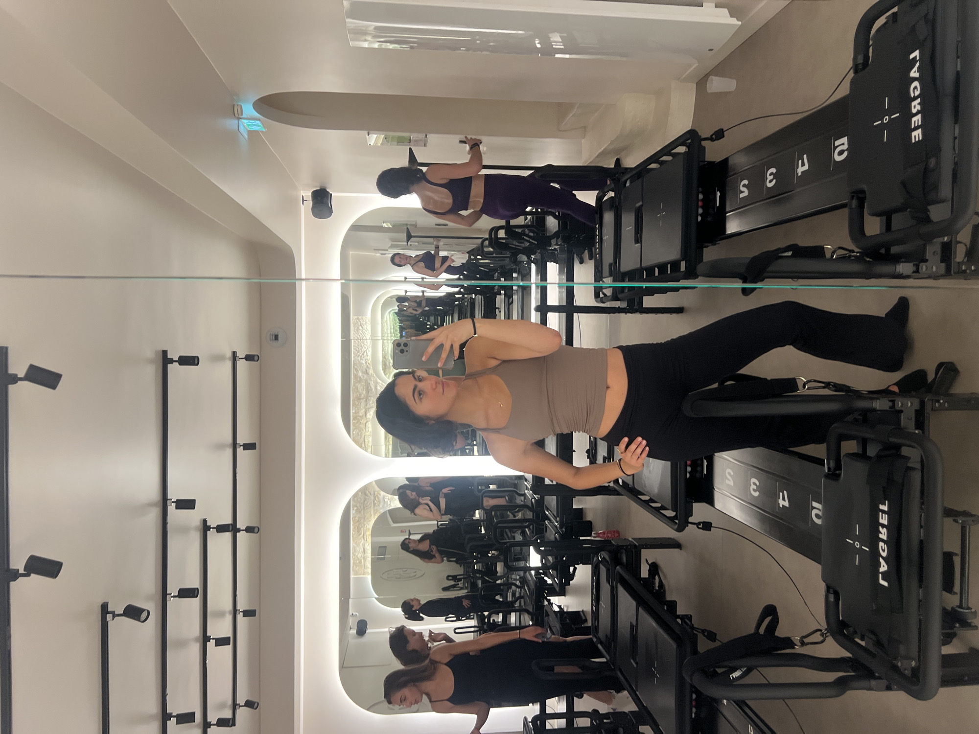 A medium-shot taken in a workout studio is reflected in a large mirror extending the length of the studio's wall. A young woman, presumably the photographer, is taking a selfie with her phone while positioned on a pilates reformer machine. She is wearing a tan tank top and black leggings. Behind her, the reflection shows several other people participating in pilates exercises on reformer machines. The studio has white walls with a series of black track lighting fixtures on one wall. The reformer machines are black with white lettering. The overall ambiance is bright and modern. The location is likely in Paris, France due to the architectural style commonly found in the city.