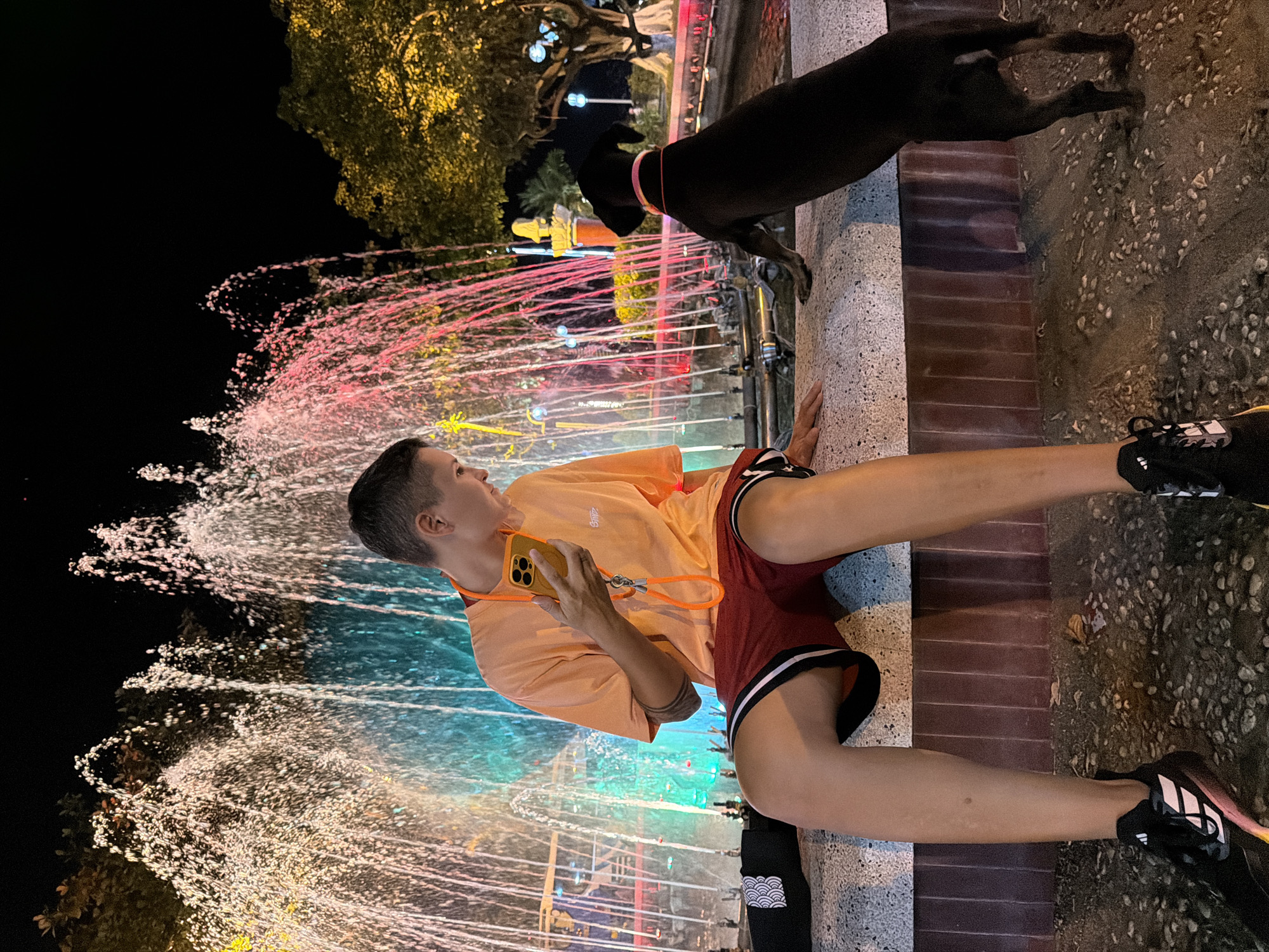 A young person sits on a stone ledge in front of a brightly lit fountain at night. They are wearing an orange t-shirt and red shorts, and holding a yellow phone. A black dog, wearing a pink collar, stands on the ledge next to them, looking towards the fountain. The fountain sprays water in various colors, including pink, blue, and yellow, and is illuminated by lights. Trees are visible in the background, also lit by yellow lights. The ground in front of the ledge is covered with gravel. The scene appears to be in a public park or plaza in Nha Trang Ward, Vietnam.