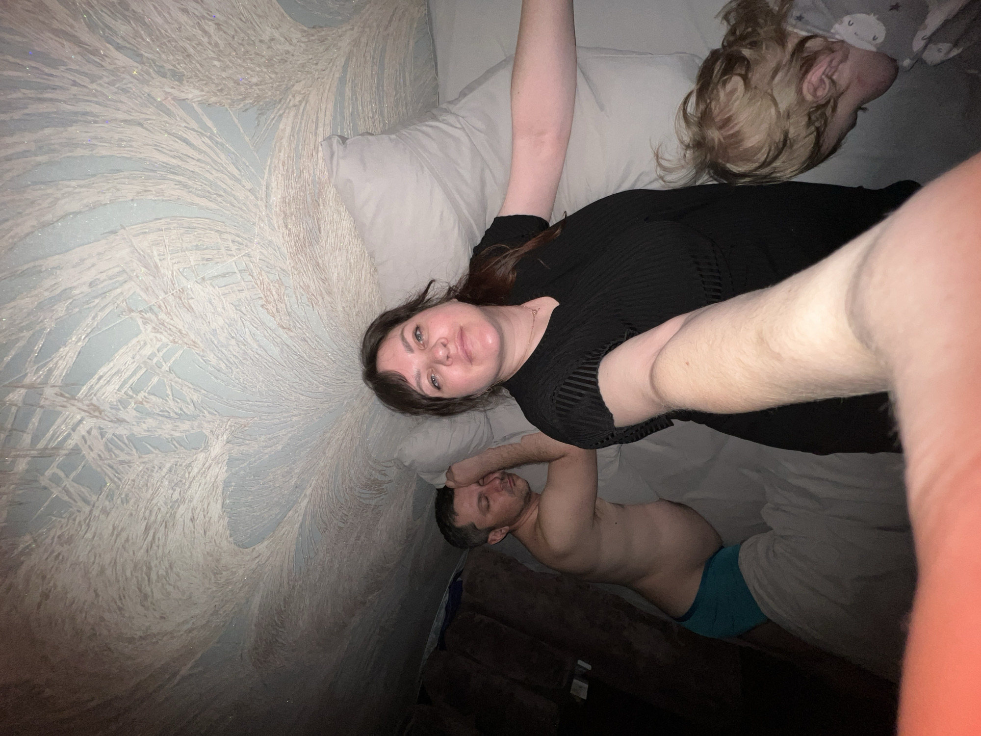 A woman in a black dress is lying in bed next to a man in blue underwear. A child is lying on a pillow behind the woman. The wallpaper has a feather pattern. The location is an interior. The city of Noviy, Russia is not visually apparent in the image.