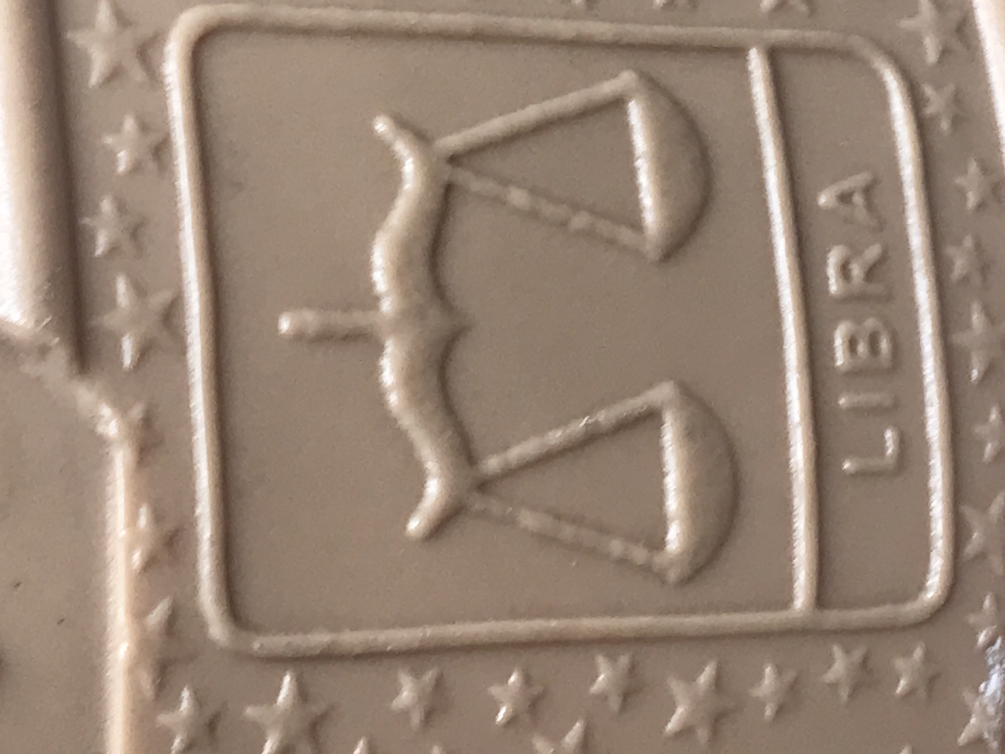 This close-up image displays a light brown or beige textured surface, likely made of molded plastic or rubber. The primary subject is an embossed design featuring the astrological symbol for Libra, which is a set of weighing scales, centered within a rectangular border with rounded corners. The scales symbol is rendered in relief, showing distinct raised lines and shapes.
To the right of the scales symbol, within the same rectangular frame, the word "LIBRA" is embossed vertically, with letters stacked from top to bottom. Surrounding this central rectangular design, numerous small, five-pointed stars are embossed into the surface, forming a decorative border. The lighting is soft and diffused, creating subtle shadows that emphasize the three-dimensional quality of the raised elements. The surface itself appears to have a slightly grainy or matte texture, while the raised parts catch the light, showing a smooth finish.
No people or activities are present in the image, which focuses solely on the details of this molded object. While the location context of Onitsha, Nigeria is provided, there are no visual cues within the image itself to suggest this specific geographical setting. The design is generic and could be found on various manufactured items, such as the sole of a shoe, a mat, or a decorative plaque. The only visible text is the word "LIBRA."