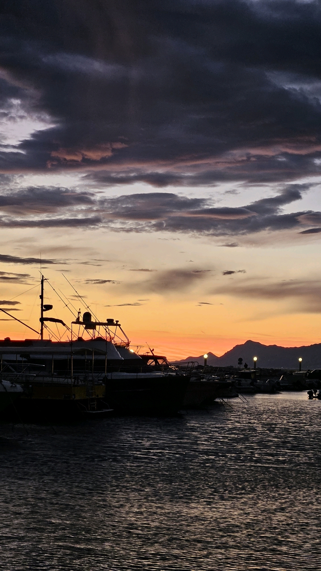 The image captures a harbor scene at dusk in Općina Tučepi, Croatia. In the foreground, dark, rippling water fills the lower portion of the frame, reflecting the muted colors of the sky. Silhouetted against the horizon are several boats moored in the harbor. One prominent boat in the mid-ground features a tall mast with antennae and various rigging. Behind the boats, a range of mountains forms the distant background. The sky is a dramatic gradient of colors, transitioning from deep purples and grays of the clouds at the top to vibrant oranges and yellows near the horizon, indicating the sun has recently set. Several lights are visible along the waterfront, adding small points of illumination to the darkening scene. There are no people visible in the image.