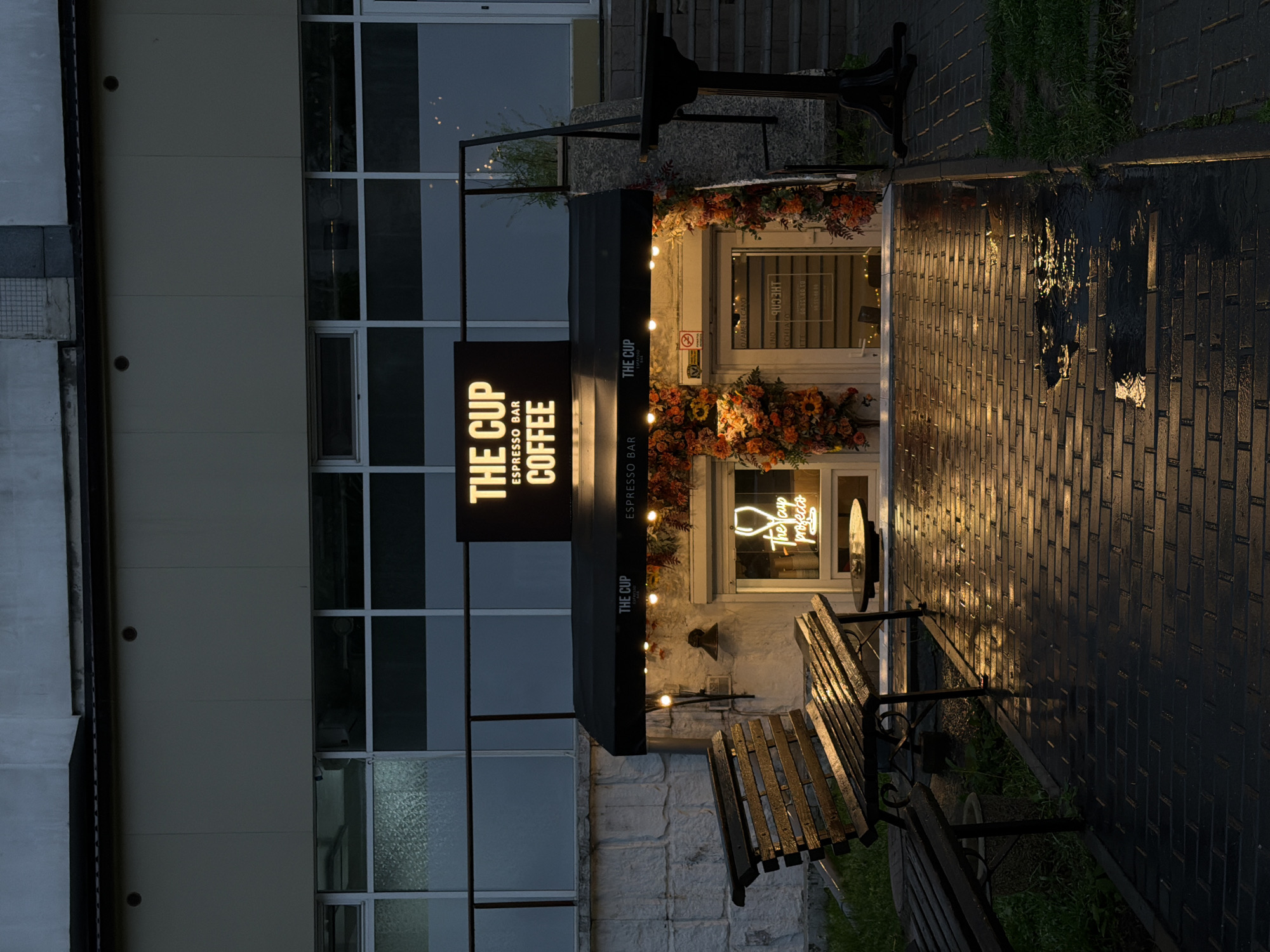 The image depicts the facade of "The Cup" coffee shop, located in an urban setting of Unknown, Unknown. The shop's entrance is decorated with autumnal flowers, and a black awning shades the doorway. A sign vertically displays the shop's name and offerings in bright yellow lettering. A bench sits adjacent to the shop's entrance on a wet, brick sidewalk reflecting the warm light emanating from within the shop. The building adjacent to the coffee shop is multi-story, with large pane windows visible, and lacks any immediate identifying markings. A vintage clock-like circular sign is attached near the window.
