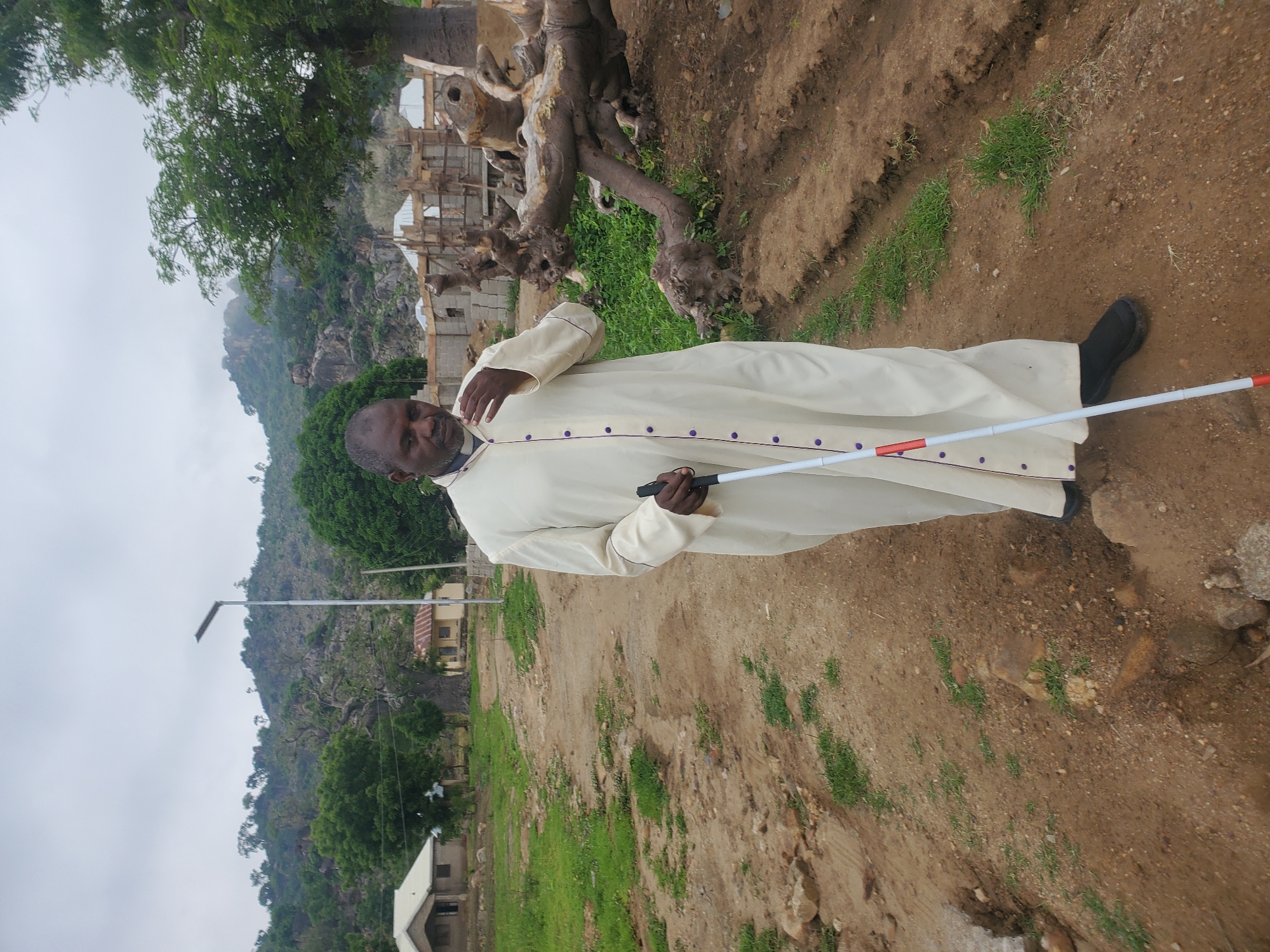 A man wearing a cream-colored robe with purple buttons stands on a dirt path. He is holding a white cane with red and white markings. His right hand is raised to his face, as if he is speaking or thinking. The background shows a rocky, green landscape with trees and some simple buildings. A solar-powered lamp post is visible on the left side of the frame. The sky is overcast.
