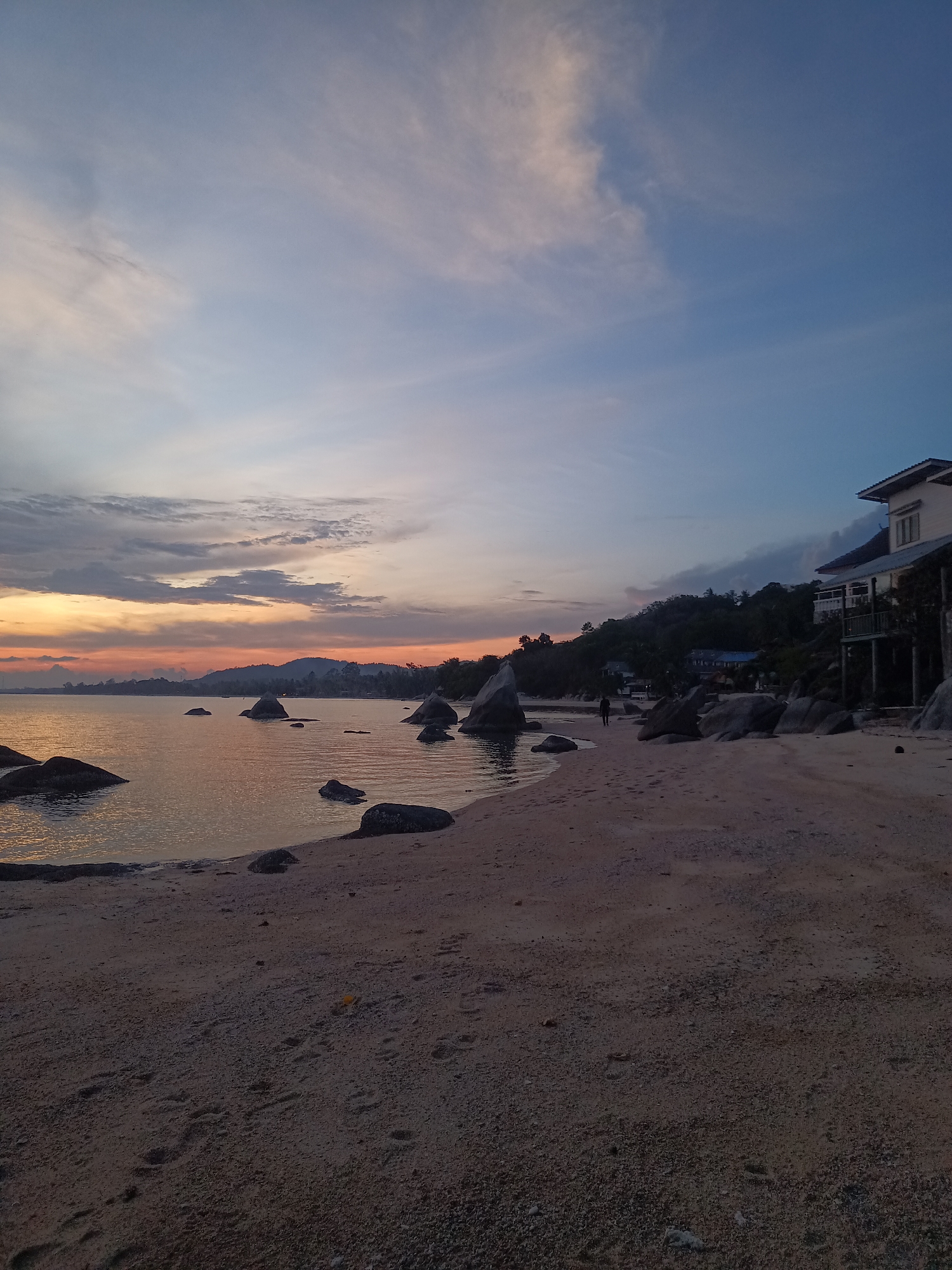 The image shows a beach at dusk in Baan Kai, Thailand. The sky is a mix of orange, yellow, and blue hues, indicating a recent sunset. Several rocks are scattered along the shoreline and in the water. In the background, there is a building on a small hill, and some vegetation. There appears to be one person walking along the beach. The sand in the foreground is textured, and it shows footprints. The sea is calm, reflecting the colors of the sky.