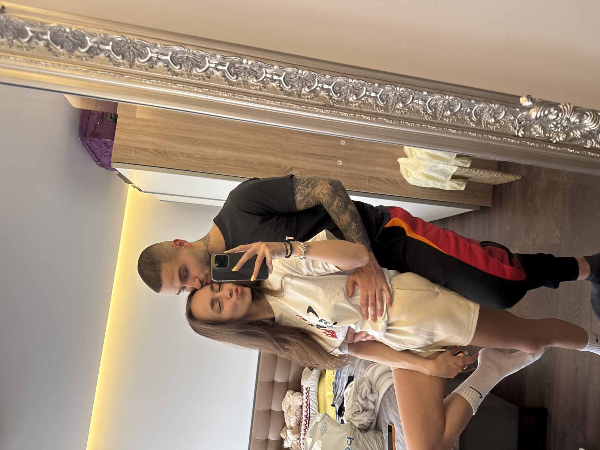 A man and a woman are taking a selfie in a mirror indoors. The man is bald, wearing a black shirt, black and red sweatpants, and has tattoos on his arms and neck. He is kissing the woman's forehead while holding a phone in his hand. The woman has long brown hair and is wearing a white t-shirt, white shorts, and white socks. They are standing in what appears to be a bedroom, with a bed and wardrobe visible in the background. The image is a reflection in a mirror with a decorative silver frame. The location appears to be indoors, based on the objects present in the room. It is impossible to determine the exact location in Sofia, Bulgaria, from the image.