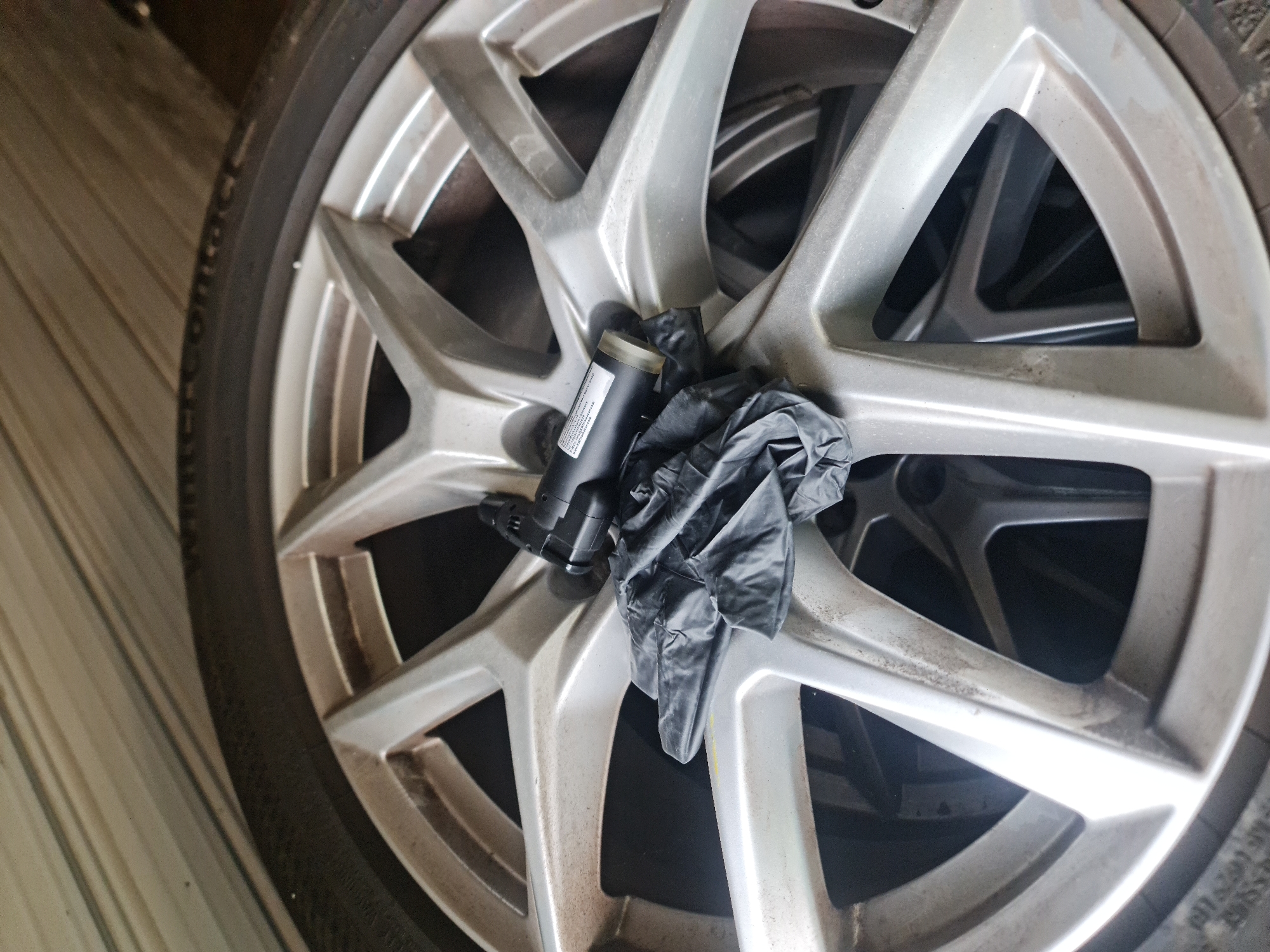 A close-up, slightly high-angle shot captures a car wheel with a silver multi-spoke alloy rim and a black tire, positioned in what appears to be a garage or storage setting in Buzovgrad, Bulgaria. Another wheel is partially visible directly behind it. Resting on the center of the visible wheel rim is a black plastic component, possibly a car part like a washer fluid pump, alongside a crumpled black disposable glove.
The wheel rim shows signs of dirt or dust accumulation, indicating it may not be freshly cleaned or is in storage. The tire's sidewall has raised lettering, with "CONTACT" being partially legible, possibly referring to a "Continental" brand tire. Other faint text includes "RADIAL" and "TUBELESS".
The black plastic component has a white label with clearly visible text: "MADE IN FRANCE", "VALEO", and part numbers or specifications like "210410" and "12V N=418919". A QR code is also present on the label, along with the website address "VALEO.COM".
The background behind the wheel consists of light-colored, possibly horizontally paneled or grooved surfaces. There are no people visible, and no activity is taking place; the items are static. The lighting suggests an indoor or shaded environment.