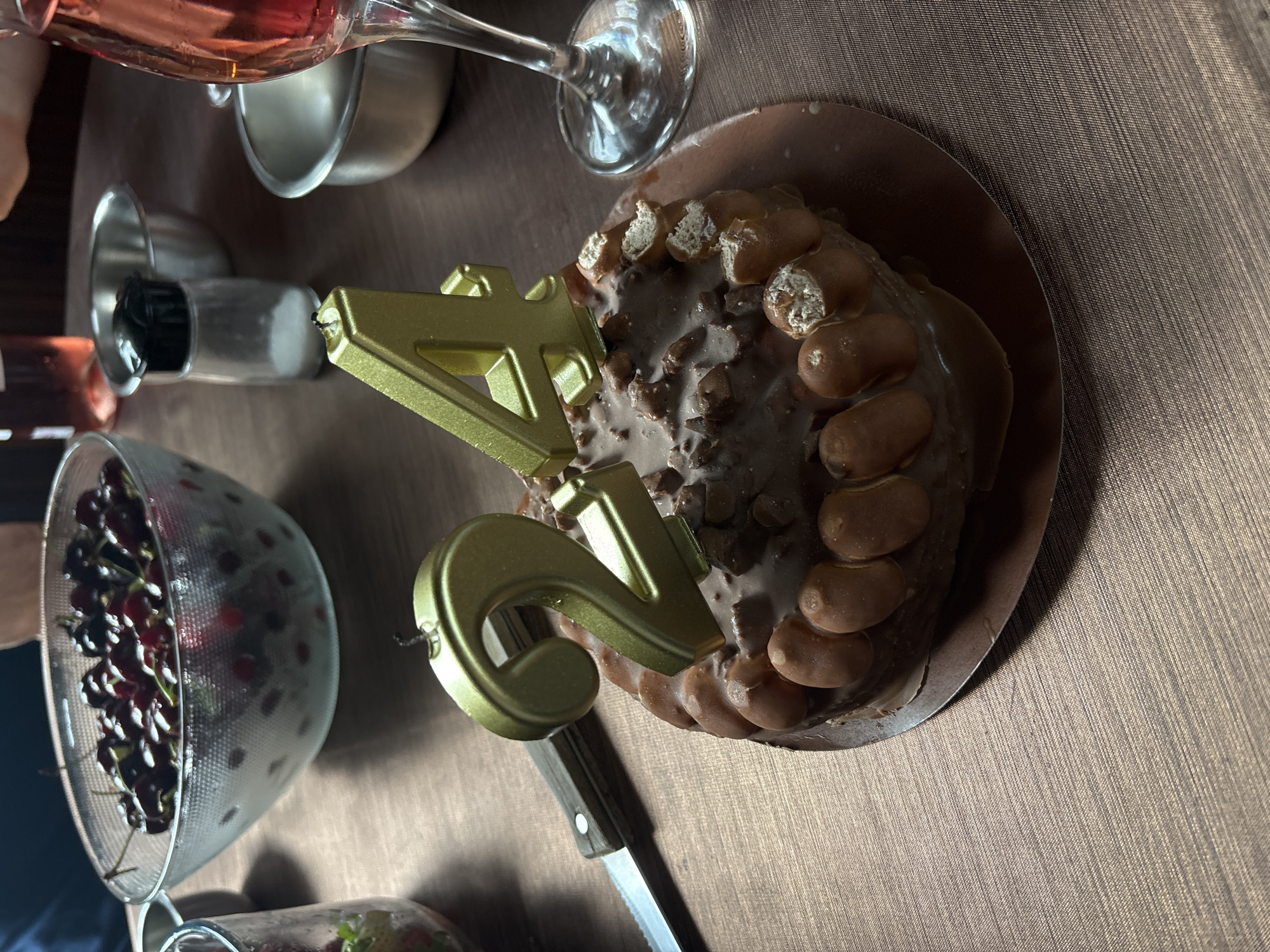 The image depicts a celebratory scene on a brown tablecloth. A chocolate cake, decorated with small, filled pastries and topped with gold-colored number candles forming "42", sits on a brown plate. A serrated knife is positioned next to the cake.
A bowl filled with cherries is visible on the left side of the image. Adjacent to the bowl are a bottle and a salt shaker. In the top left corner, there is a glass of pink liquid, possibly rosé wine or juice. The background suggests an indoor setting.
There is no information present in this image that allows it to be correlated with the city of Varna, Bulgaria.