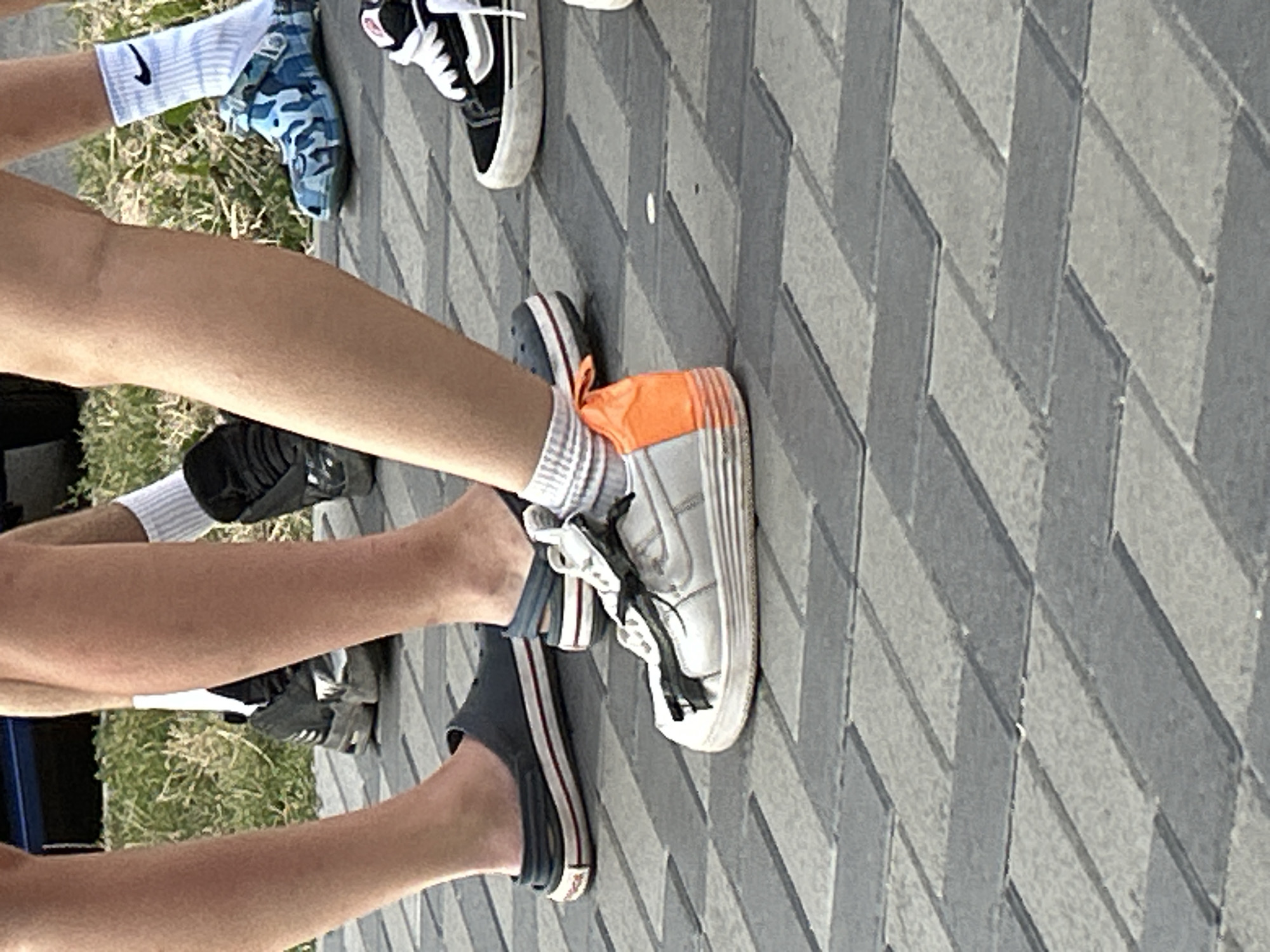 The image shows a close-up view of several pairs of feet and legs standing on a patterned walkway. The walkway is made of rectangular paving stones laid in a herringbone pattern, alternating between light grey and darker grey. In the background, there is a patch of green grass with some dry, yellowed vegetation.
The people are wearing various footwear. On the left, a person is wearing dark grey slip-on shoes with a red and white striped sole. Next to them, another person is wearing white sneakers with black laces and a prominent orange heel accent. The laces on these sneakers are tied in a bow and appear to be black.
To the right, one individual is wearing blue and white camouflage Crocs with a Nike logo on the white sock. Further to the right, a person is wearing black Vans sneakers with white side stripes and white laces. The socks worn by this individual are white with a Nike logo.
The image is a medium shot, focusing on the lower legs and feet of the individuals. The lighting suggests it is daytime, with soft shadows indicating an overcast or partially shaded environment. The overall impression is of a casual gathering or a group of people walking outdoors. The specific location in the city of Brovary, Ukraine, cannot be determined from the visual information alone.