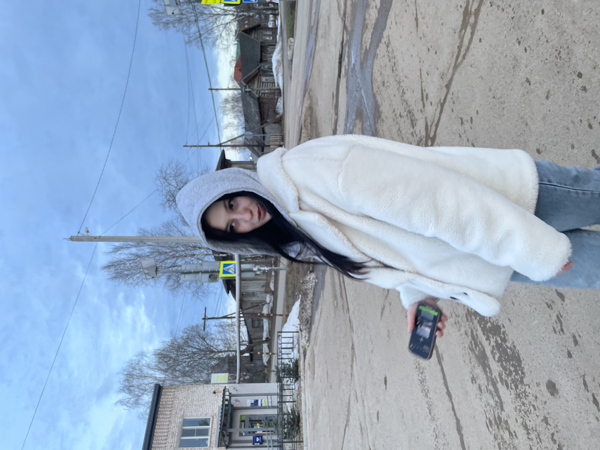 A young woman is standing on a concrete road. She is wearing jeans and a white fleece jacket with a gray hood. She is holding a black smartphone. In the background, there are buildings and utility poles. The location is in Фоки, Russia.