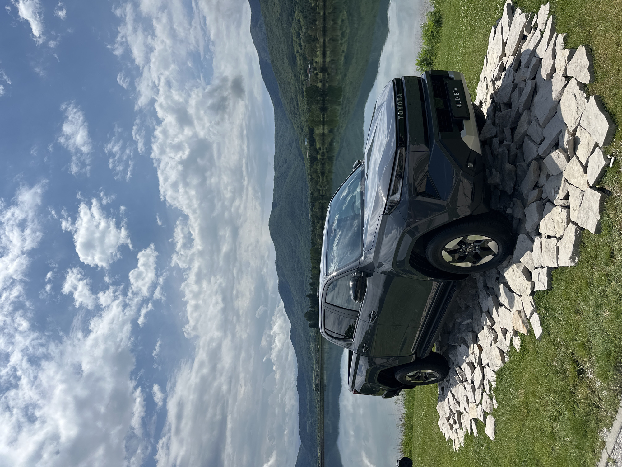 The image captures a dark grey Toyota Hilux pickup truck positioned on a grassy bank next to a calm body of water, likely a lake or wide river, in Razliv, Bulgaria. The truck is angled slightly towards the viewer's right, showcasing its front.
In the background, across the water, a range of green, forested mountains extends horizontally, with their peaks clearly reflected on the water's still surface. Above the mountains and water, the sky is a vibrant blue dotted with scattered white cumulus clouds, indicating bright, fair weather.
In the foreground, around the truck's front section, a considerable mound of large, irregularly shaped, light-colored rocks is arranged on the grass, possibly as part of a display or to demonstrate the vehicle's ruggedness. Sunlight casts clear shadows from the truck and the rocks.
Visible text on the vehicle includes "TOYOTA" emblazoned in white on the front grille. Additionally, a rectangular sign located near the front bumper prominently displays "HILUX BEV" in white text, indicating that the vehicle is a Battery Electric Vehicle. No people are visible in the scene. The overall impression is a display or showcase of the electric pickup truck within a picturesque, mountainous, and watery natural environment.