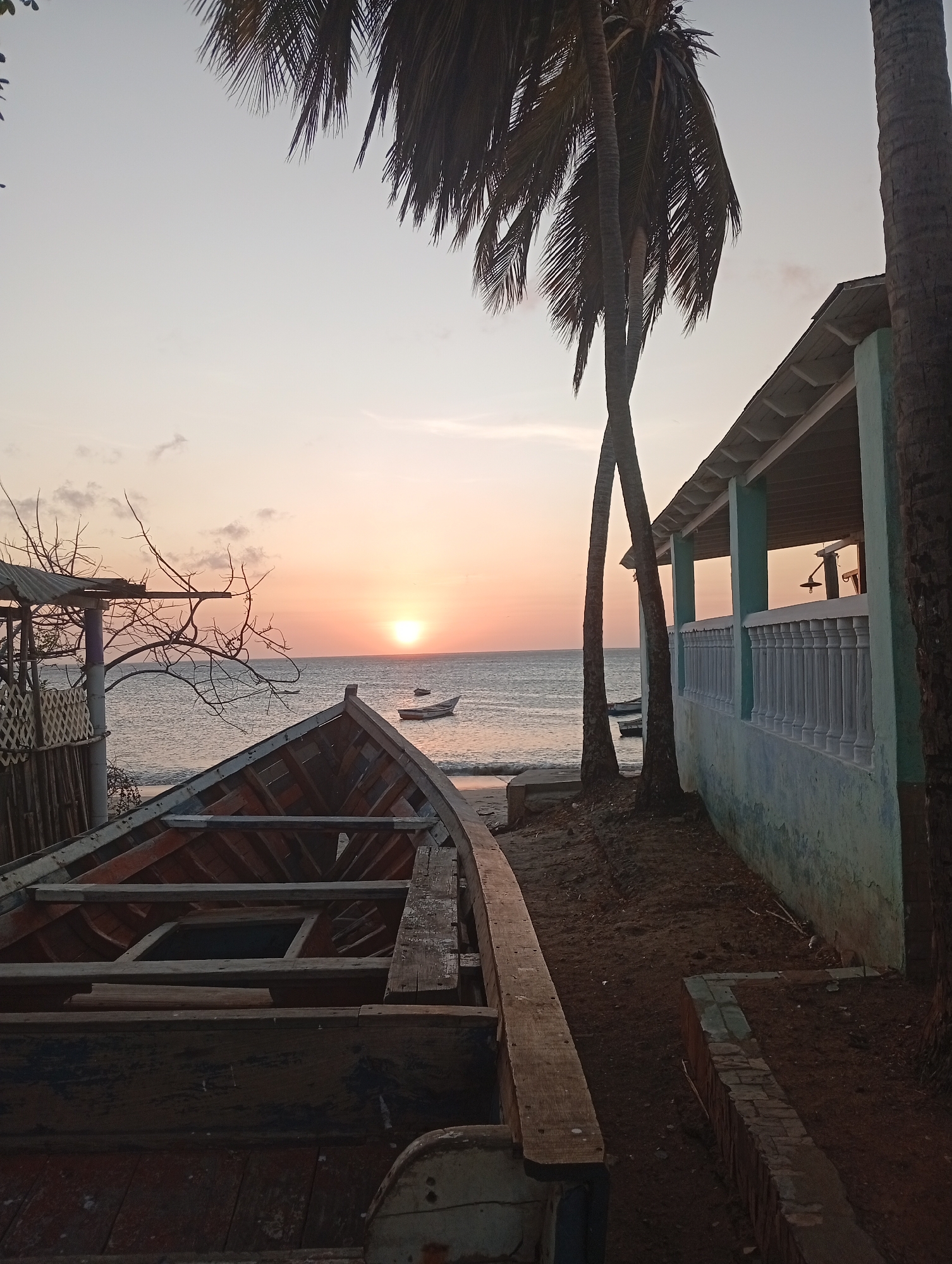 The image depicts a coastal scene in Juan Griego, Venezuela, at sunset. In the foreground, there is an old, wooden boat resting on the shore. To the right, a light blue building with a white balustrade is visible. Palm trees frame the building. In the background, the sun is setting over the ocean, with a small boat silhouetted on the horizon. The sand is a dark, earthy color, and the sky is a gradient of orange and pink hues.