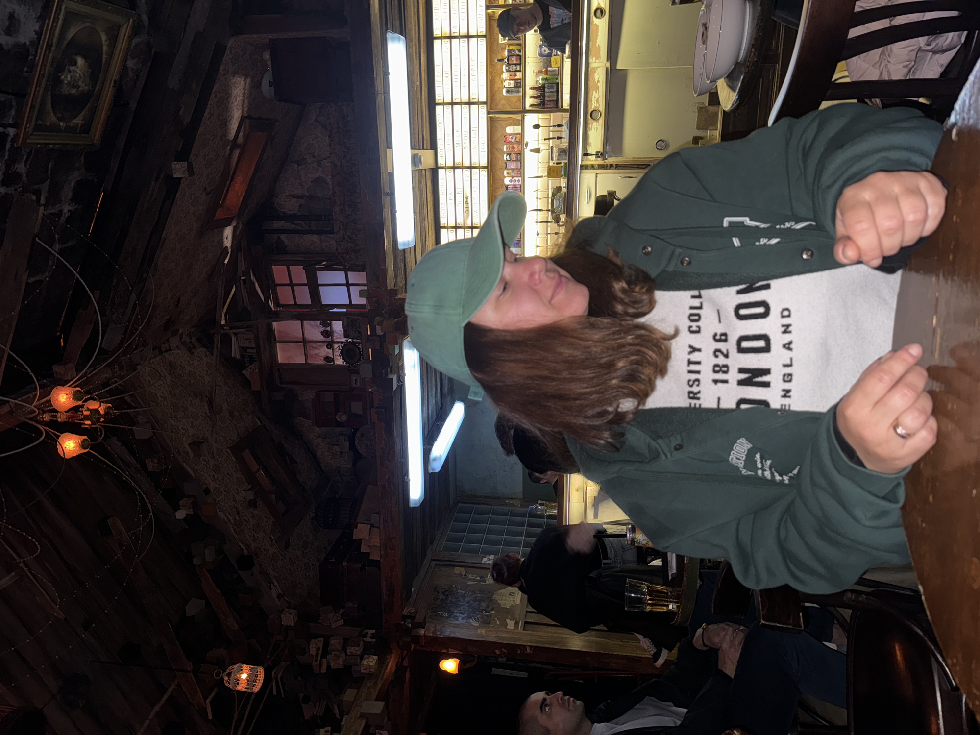The image shows the interior of a bar in Moscow, Russia. The bar has a rustic decor with wooden beams, exposed brick, and vintage light fixtures. A woman wearing a green baseball cap and a sweatshirt with "London" printed on it sits at a wooden table. Other patrons are visible in the background, some seated at the bar. The bar appears to be well-lit with fluorescent lights and decorative lamps. A menu board listing various drinks is visible behind the bar.