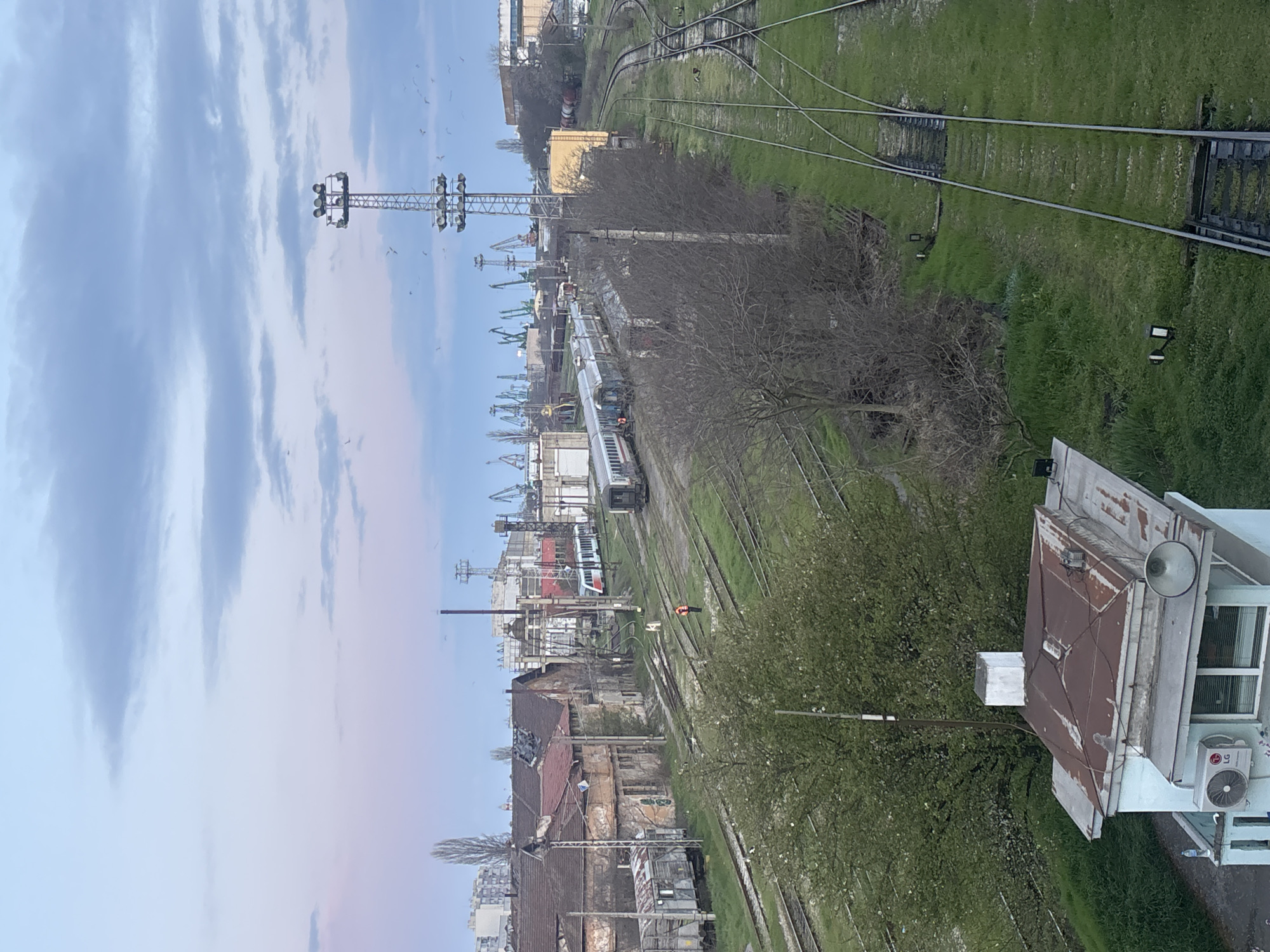 This photograph, taken in Varna, Bulgaria, shows a railway yard with multiple tracks. A train is positioned on one of the tracks, composed of silver passenger cars, stopped near a person in a traffic vest. In the background, harbor cranes are visible. Dilapidated buildings are located on the left-hand side of the image, while greenery occupies the right-hand side. A rooftop of a small building is prominent in the foreground. In the distance, a floodlight tower is visible beneath a partially cloudy sky. Birds are also visible in the sky.