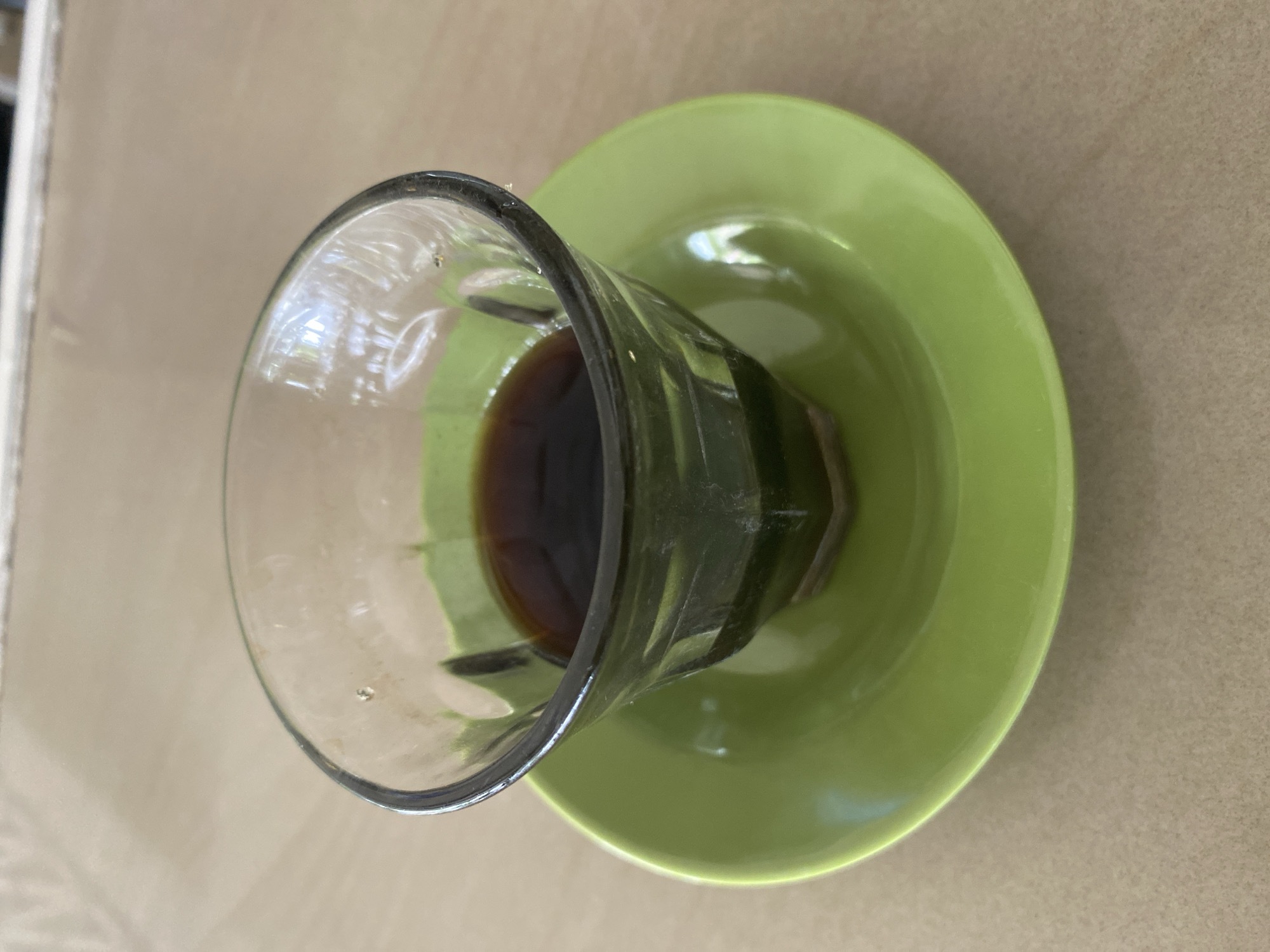 The image shows a glass of coffee on a green saucer, viewed from directly above. The coffee is dark brown and partially consumed. The glass is clear, with a distinct rim and ribbed design. The saucer is plain green. The table has a light brown surface. The location is Lhokseumawe, Indonesia.