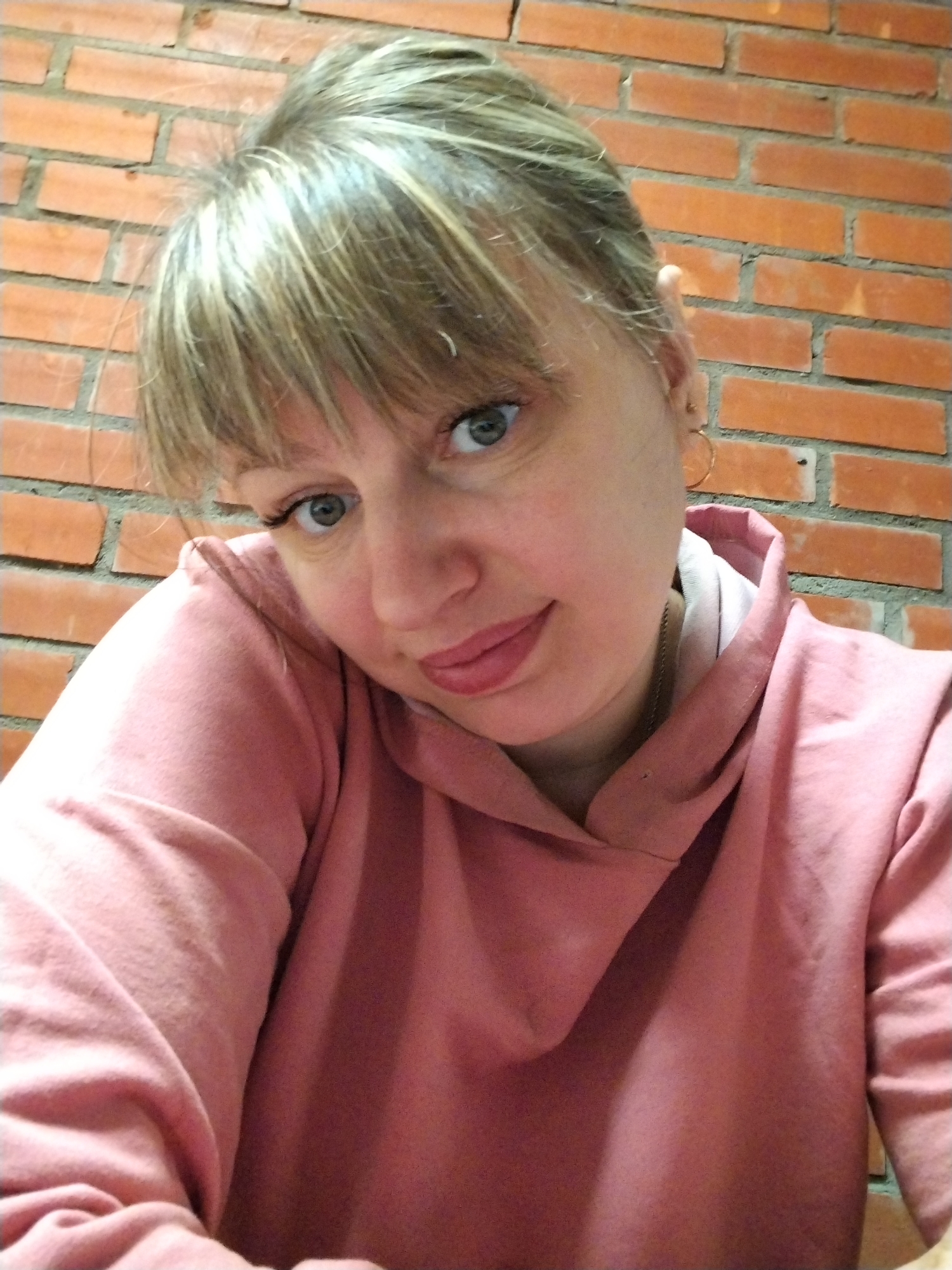 A close-up portrait shot features a woman looking directly at the camera. She has blonde or light brown hair styled with bangs across her forehead and light blue or green eyes. Her expression is soft and slightly inquisitive, with her lips parted slightly, suggesting a gentle smile. She is wearing a dusty rose-colored hooded sweatshirt. A faint gold chain is visible around her neck, and a small gold hoop earring adorns her right ear. The background consists of a rustic red-orange brick wall with grey mortar, implying an indoor setting or a covered outdoor area. The lighting is soft and even, suggesting ambient indoor light. The photograph captures a serene and personal moment in Шебунино, Russia. No visible text is present.