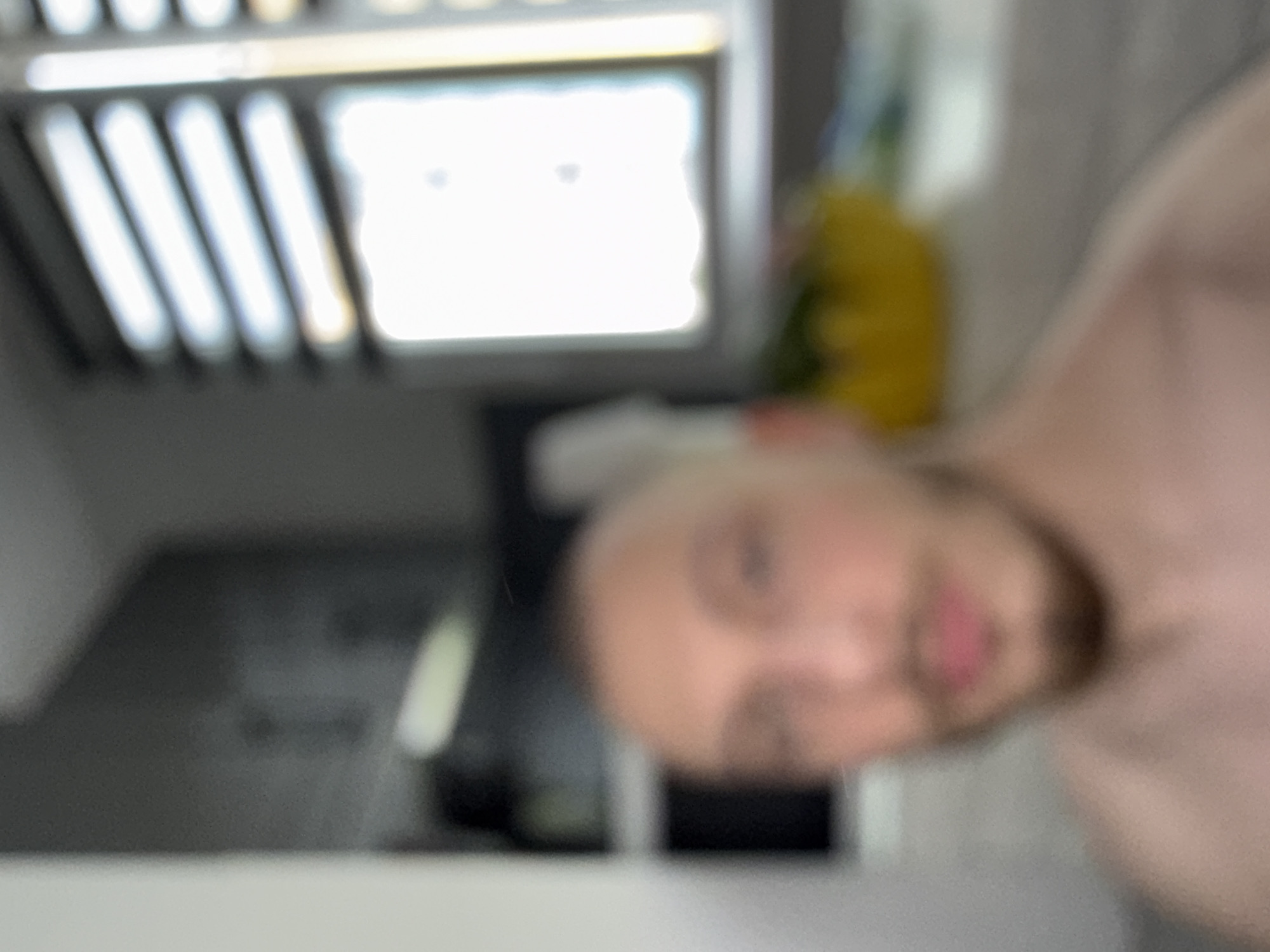 The image is heavily blurred, making precise identification difficult. The primary subject appears to be a bald man with a light complexion and a visible beard, possibly facing the camera. Behind him, an out-of-focus kitchen setting can be inferred, including a rack with parallel metal bars, a window-like bright light source, and a yellow object. The specific location in Stara Zagora, Bulgaria, cannot be determined due to the poor image quality and lack of discernible landmarks.