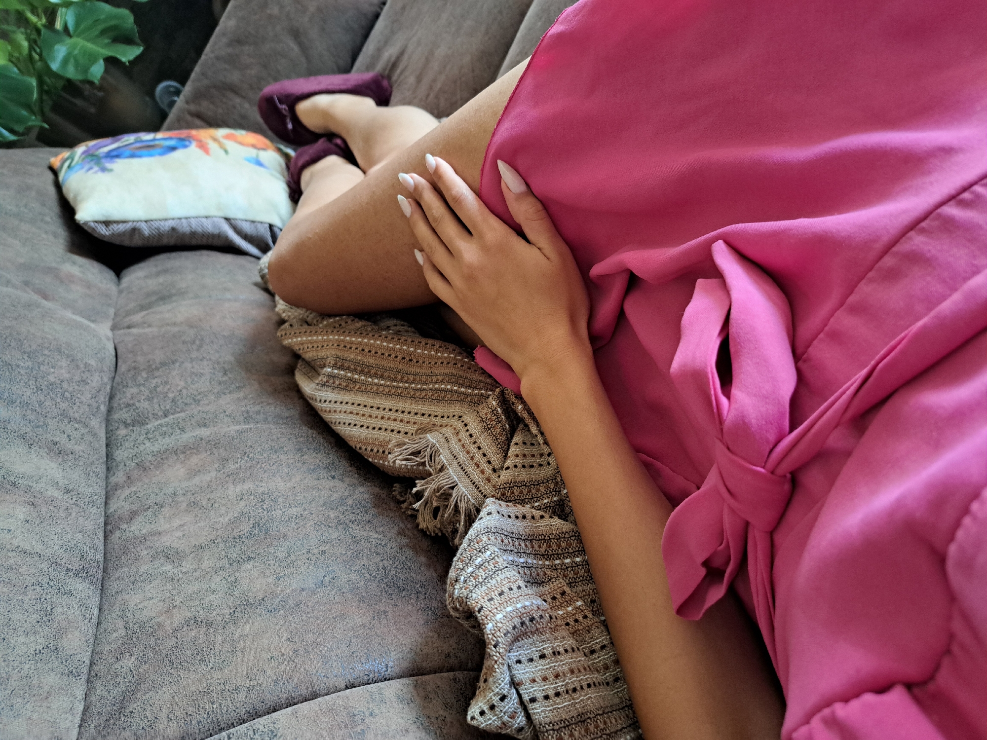 A person is lying down on a brown sofa, with their legs crossed and a hand resting on their thigh. The person is wearing a bright pink garment that appears to be a dress or skirt, tied at the front with a bow. A patterned throw blanket is draped over the person's legs. A decorative pillow with a colorful floral design is visible behind the person's legs. The environment appears to be indoors, likely a living room, with a plant visible in the background. The lighting suggests it is daytime.