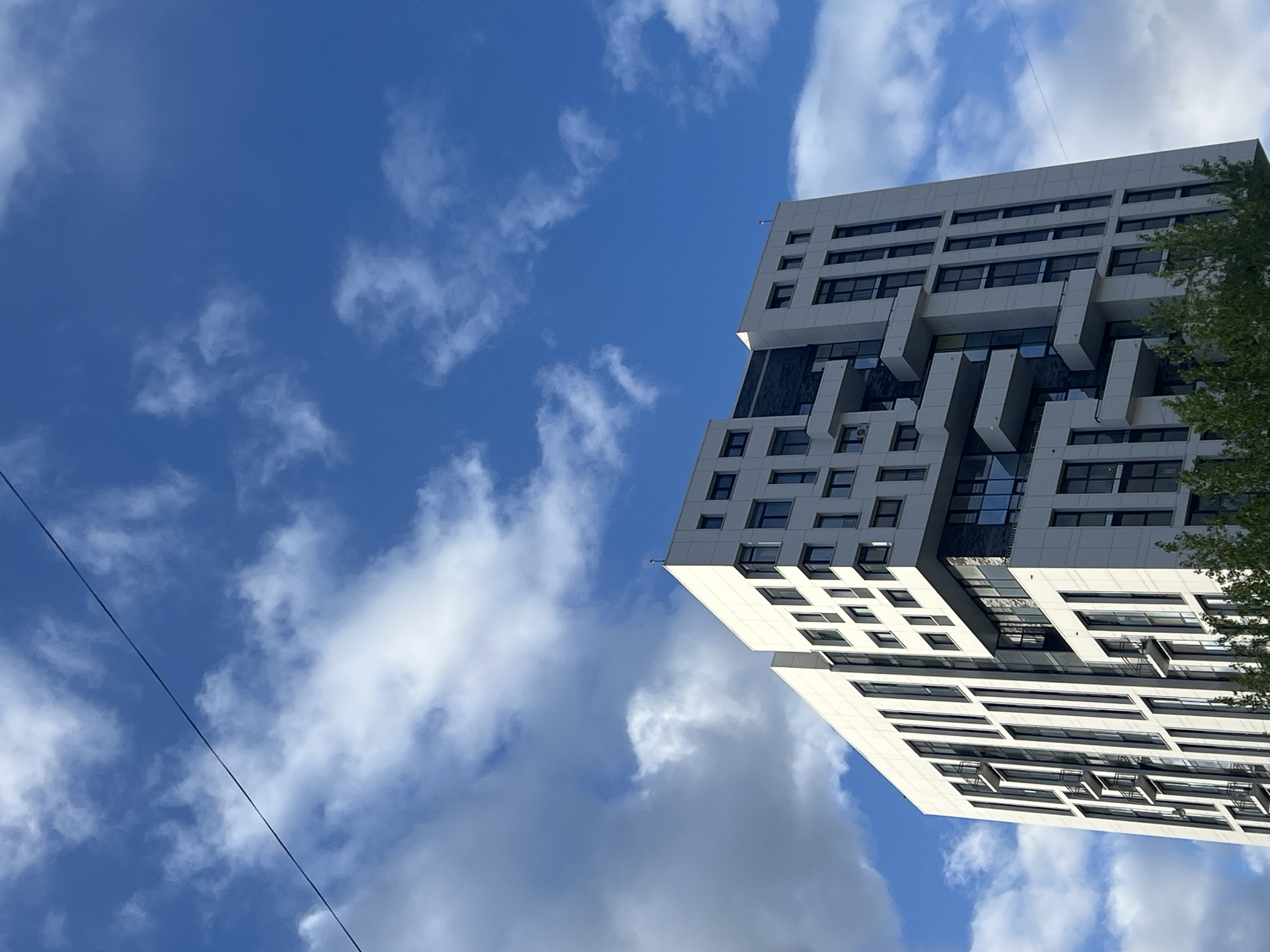 The image shows a view looking up at a modern, multi-story building in what appears to be the city of Perm, Russia, as indicated by the architectural style. The building has a white facade with dark windows and a stepped, geometric design. The sky is blue with scattered white clouds. A thin power line is visible diagonally across the lower left portion of the image. The building is located in the city of Perm, Russia, the exact location is not specified in the given images.