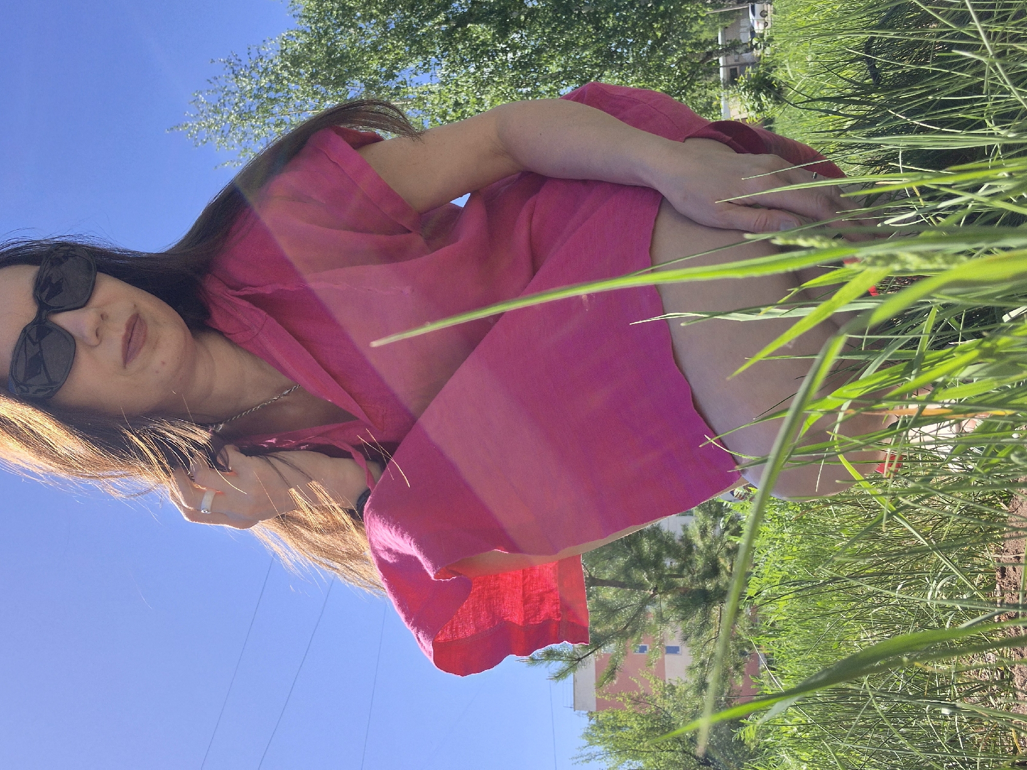 A low-angle, outdoor shot shows a woman in a bright pink dress squatting down near a patch of tall, green grass. She is wearing sunglasses and has long, dark hair that cascades over her shoulder. Her skin tone is fair. The sky is a clear, bright blue, suggesting a sunny day. In the background, one can see a building, potentially an apartment complex, partially obscured by trees. The scene is set in Unknown, Unknown.