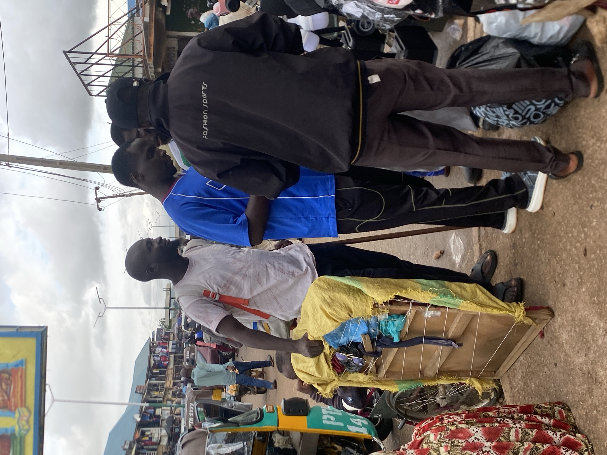 The image depicts a street scene in Bukuru, Nigeria, under an overcast sky. Three men are the central subjects. The man on the left, wearing a light pink t-shirt and dark pants, carries a wooden display stand draped with yellow and green fabric, holding various items including sunglasses and fabric scraps. He is also wearing a red backpack. To his right, a man in a bright blue t-shirt and black pants stands facing away from the camera, his head turned to his left. A third man, in a black jacket with "FASHION SPORTS" written on the back, and dark brown pants, stands with his back to the viewer, facing the other two men. Behind them, a colorful auto-rickshaw is visible, along with market stalls and other vehicles suggesting a bustling commercial area. The ground is paved with concrete and dirt. The overall impression is one of everyday commerce in a busy urban environment.
