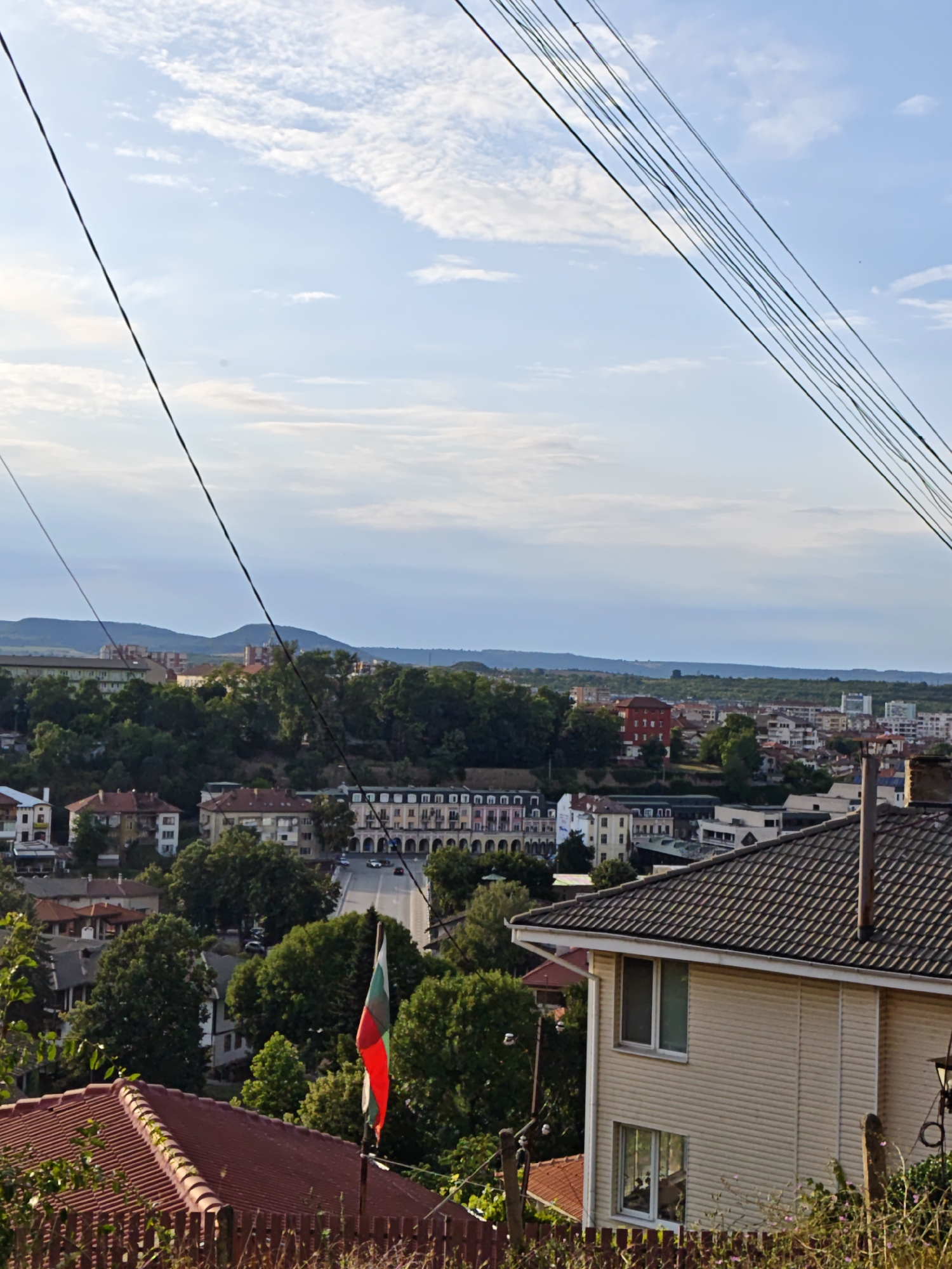 The image captures a view of the city of Lovech, Bulgaria, from an elevated perspective, likely a hillside, looking down into the urban landscape. In the foreground, a segment of a tiled roof and a wooden fence are visible. A flagpole with the Bulgarian flag, displaying white, green, and red horizontal stripes, stands prominently on the right side of the foreground.
Beyond the immediate foreground, the scene unfolds into a densely populated area characterized by numerous buildings of varying architectural styles and heights. Residential houses, some with red tiled roofs, are interspersed with larger, more modern structures. A noticeable building with a long facade featuring arched windows and balconies is situated in the middle ground. A paved road runs through the city, with a few cars visible on it, suggesting typical urban activity.
The city is nestled amongst lush green trees and foliage, indicating a verdant environment. In the distance, rolling hills form the backdrop under a sky with scattered clouds. The overall impression is that of a well-established, somewhat hilly city with a blend of old and new architecture, surrounded by nature. The presence of power lines crisscrossing the sky adds to the urban context of the image.