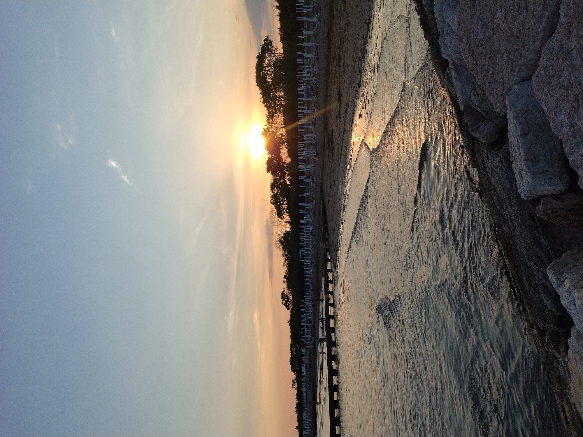 This is a vertical, eye-level shot of a beach in Caorle, Italy, at sunset. The sun, a bright orb, is partially obscured by trees on the horizon, casting a warm glow and creating a sunbeam that stretches across the scene. The sky is a gradient of pale blue at the top, transitioning to warmer hues of orange and yellow near the horizon. A few wispy clouds are visible in the sky.
In the foreground, dark, rocky sea defenses line the bottom edge of the frame, with some larger, lighter-colored rocks interspersed. Gentle waves lap against the shore, reflecting the sunset's light. A wooden pier, supported by evenly spaced pylons, extends into the water from the left side of the image. Several small figures, likely people, are visible on the pier, some appearing to walk or stand.
The beach itself is sandy, with rows of white beach chairs and umbrellas set up, suggesting a developed tourist area. More figures are scattered across the beach, some near the water's edge and others further back. A line of trees forms a dense backdrop behind the beach chairs, silhouetted against the sunset. The overall atmosphere is serene and peaceful, characteristic of a beach sunset.