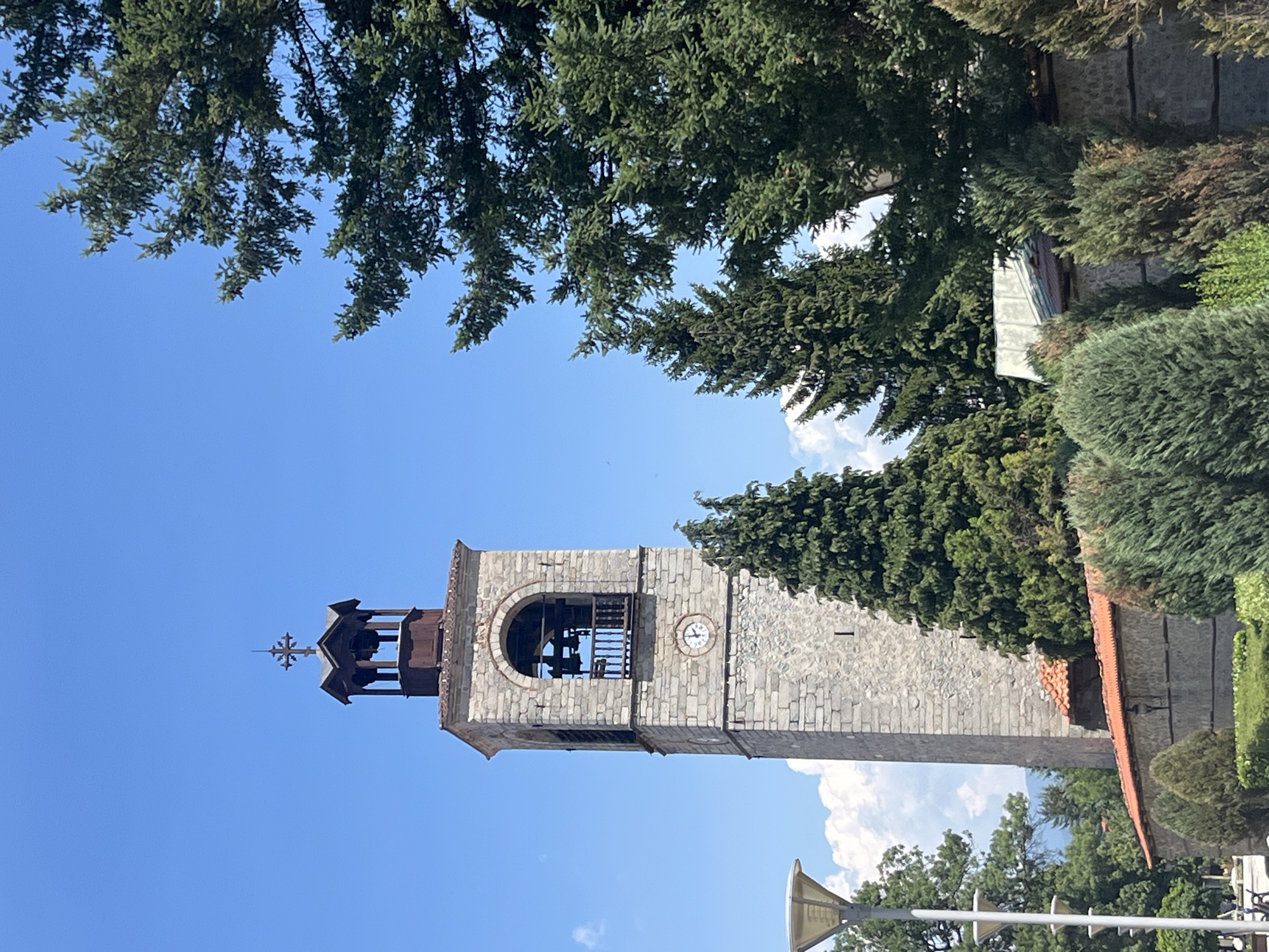 In Bansko, Bulgaria, on a clear day with a bright blue sky dotted with a few white clouds, a stone clock tower stands tall. The tower, constructed from weathered grey stones, features an arched opening near its top, housing bells and a wooden balcony. A clock face with Roman numerals is visible on one side of the tower. The tower is topped with a dark, octagonal structure supporting a cross. Surrounding the tower are various evergreen trees, including tall, dense conifers and smaller, rounded bushes. A low stone wall with a tiled roof runs in front of the tower, partially obscuring the lower portion of the structure. In the foreground on the left, a modern lamppost with a wide, conical shade is visible. The overall scene depicts a historic landmark within a green, possibly park-like, environment.