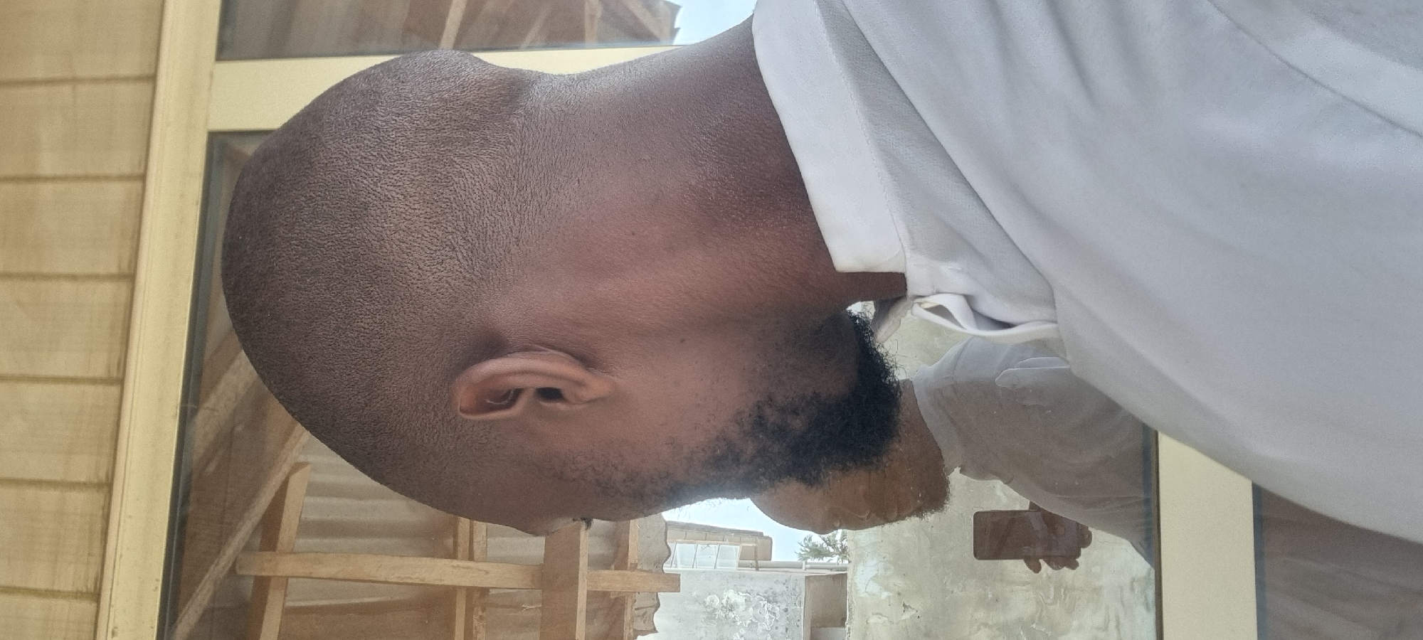 A man with a dark complexion and a short haircut, wearing a white shirt, stands beside a window. The window frame appears to be made of light-colored wood. Through the window, a view of a building with a corrugated roof structure and a partial view of the sky can be seen. The man has a beard. The reflection in the window shows the man holding a dark object, possibly a phone. The setting suggests an urban environment. Given the prompt context, this could possibly be Lagos, Nigeria.