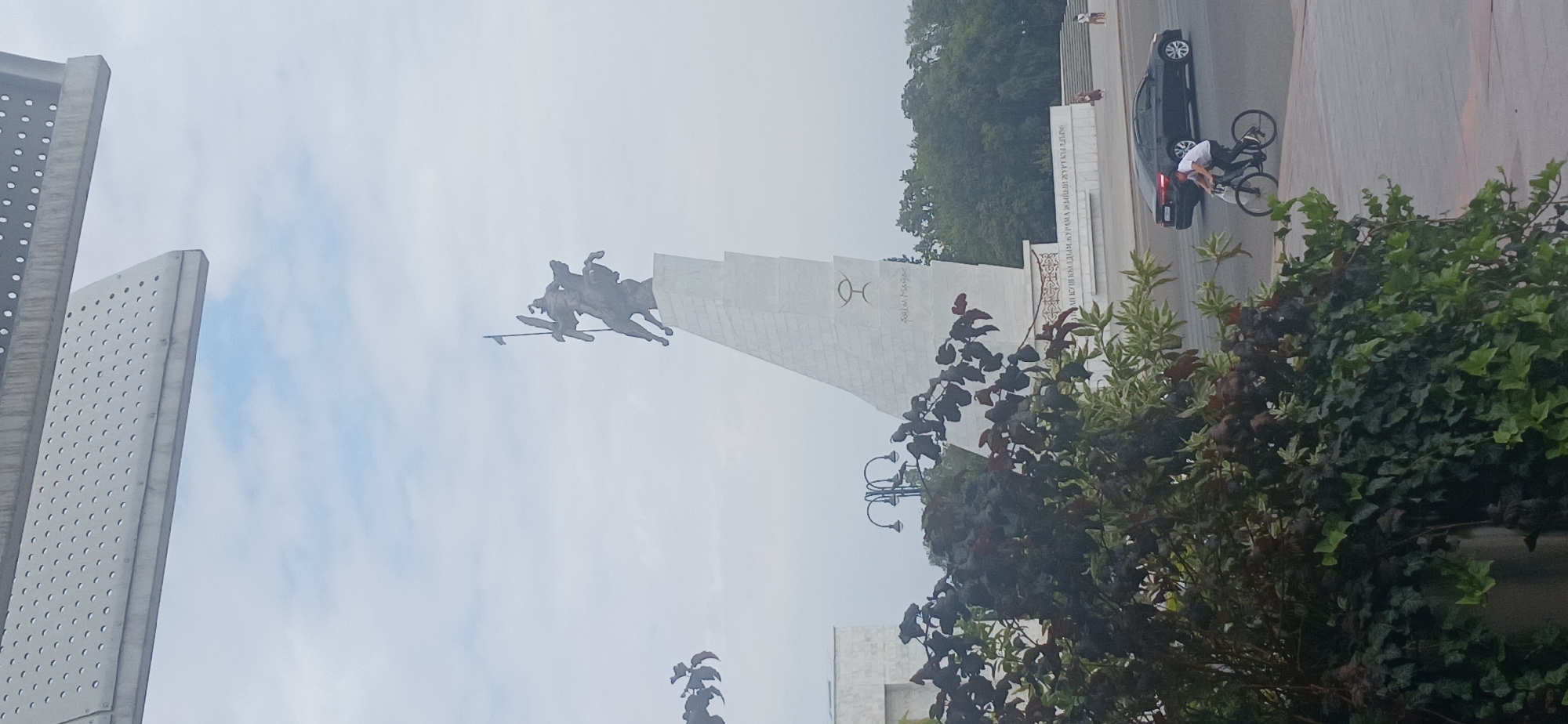 The image shows a monument of two figures on horseback, one holding a spear, atop a large, angular stone structure. The monument is situated in an open, paved area, with trees and greenery in the foreground. A black car is visible on the right, and a person is riding a bicycle past it. Several other individuals are present in the background, some walking near a set of stairs. The sky is overcast with clouds. The monument is the Monument to the Heroes of the 1916 Central Asian Uprising located in Bishkek, Kyrgyzstan, and it is often referred to as the "Kyrgyzstan" monument.