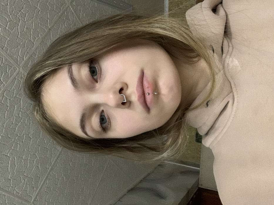 A young woman with fair skin, light brown hair, and blue eyes looks at the camera. She has a nose ring in her left nostril and a labret piercing centered below her lower lip. She wears a light beige hooded sweatshirt. The background appears to be a textured, neutral-colored wall on one side and a partially visible wooden surface on the other. The image is rotated slightly counterclockwise.