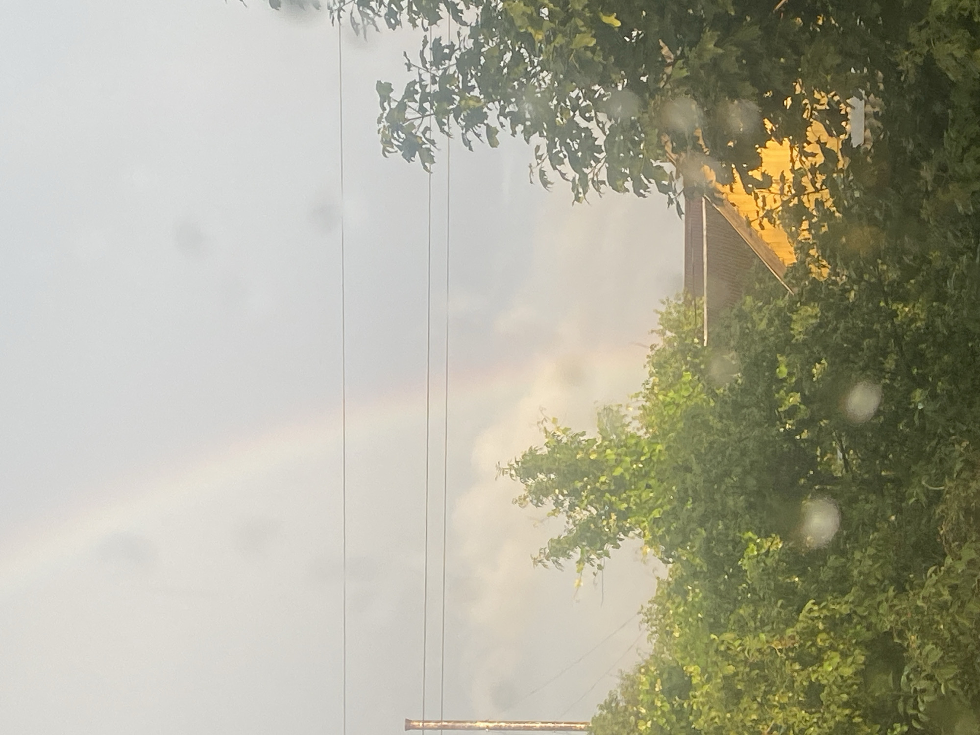 The image shows a cloudy sky with a faint rainbow visible in the upper portion. In the midground, there are several lush green trees. Partially visible behind the trees is a building with a yellow wall and a brown roof. A utility pole with wires is on the left side of the image. The scene is likely from Krupki, Belarus, given the context provided. The image appears to have water droplets on the lens or surface from which it was taken, creating blurry circular effects throughout.