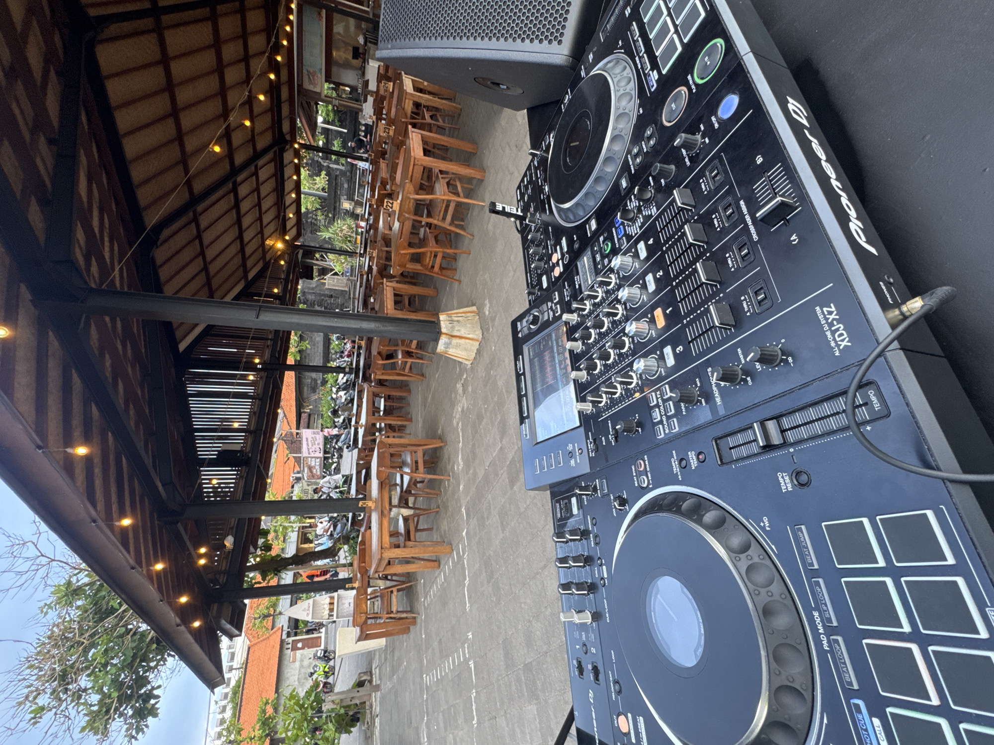 The image captures a DJ setup at an open-air venue, possibly a bar or restaurant, located in what appears to be North Kuta, Indonesia. The foreground features a Pioneer DJ XDJ-XZ all-in-one DJ system and a speaker, positioned to the right of the frame. The device's controls and branding are visible. In the background, there's an arrangement of wooden tables and chairs, typical of outdoor dining or seating areas. The venue is covered by a traditional thatched roof adorned with string lights. Further in the background, one can see buildings and scooters, indicative of a residential or commercial area. The overall environment suggests a relaxed atmosphere, possibly in the late afternoon or early evening. The primary subject is the DJ equipment set against the backdrop of an Indonesian setting. No people are directly observable, though their potential presence is implied. The image focuses on setting the stage for music or entertainment within a casual, tropical environment.