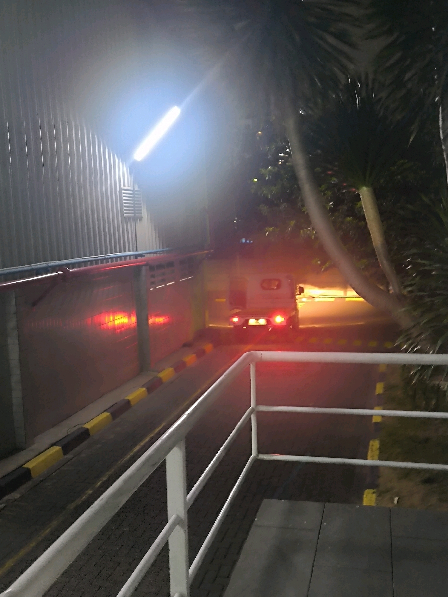 An image taken at night shows a white, three-wheeled vehicle with its brake lights illuminated, exiting a dimly lit area. The vehicle appears to be a motorized rickshaw or tuk-tuk. In the background, a corrugated metal wall is visible, lit by a bright overhead fluorescent light, casting a strong glare. To the right of the vehicle, palm trees are silhouetted against the dark sky. The ground is paved with interlocking bricks, and a yellow and black striped barrier is present along the side of the road. Red lights are reflected on the corrugated wall, possibly from the vehicle's brake lights or other sources. The scene suggests an industrial or service area due to the building material and lighting.