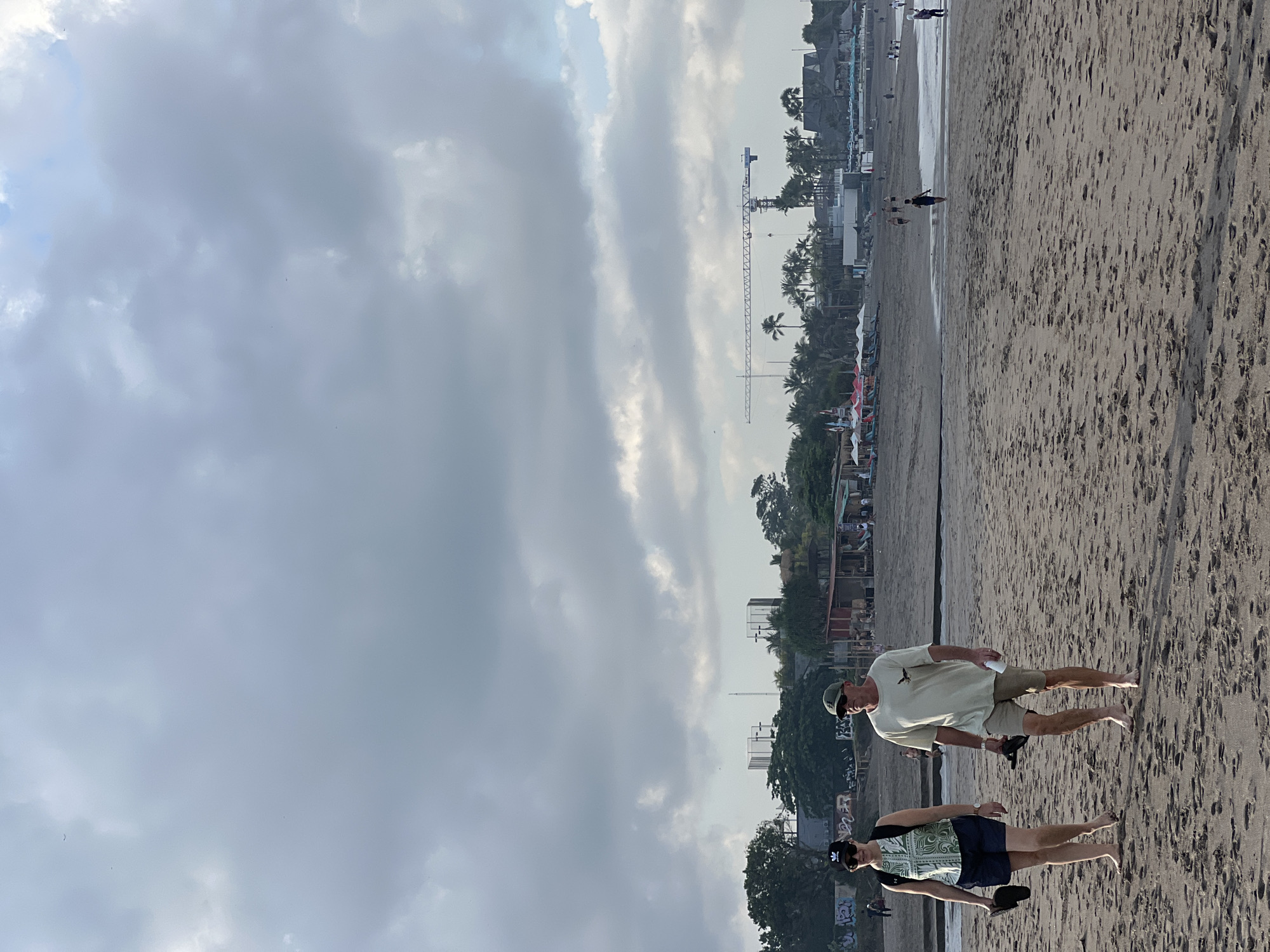 A medium shot captures two individuals walking on a sandy beach in North Kuta, Indonesia. The couple is walking towards the camera, with the woman on the left and the man on the right. The woman is wearing a patterned green and white top, dark blue shorts, and a black baseball cap. She is carrying a black bag on her back and a pair of flip-flops in her left hand. The man is wearing a light green t-shirt and khaki shorts. He is carrying a pair of flip-flops in his right hand and a white bottle in his left hand. Both are barefoot.
The beach has dark sand with visible footprints. In the background, there are several buildings, palm trees, and a construction crane. A few other people are scattered across the beach and near the buildings. The sky is overcast with gray clouds. The scene appears to be a casual day at the beach.