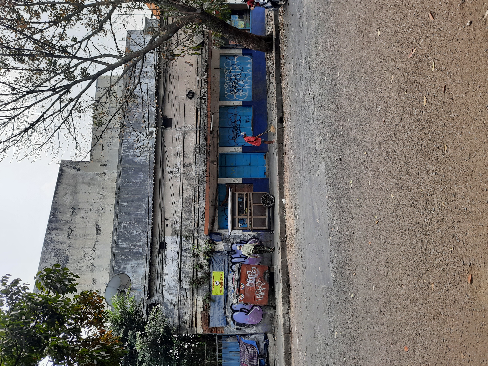 A street scene in Bandung City, Indonesia, features a weathered, graffiti-covered building with blue-shuttered windows and a blue-painted lower facade. In front of the building, a person in a light-colored shirt and camouflage pants walks away from the camera, holding a stick. To the right, another person wearing an orange shirt and a white cap sweeps the sidewalk. A small food cart with a wooden frame and a metal frame is parked on the sidewalk between the two people. A red dumpster, also covered in graffiti, is situated to the left of the food cart. The street in the foreground is paved with asphalt and scattered with dry leaves. Trees with sparse foliage are visible to the left and right of the building. Overhead wires are strung across the building. A satellite dish is mounted on the upper left section of the building. The sky is overcast.