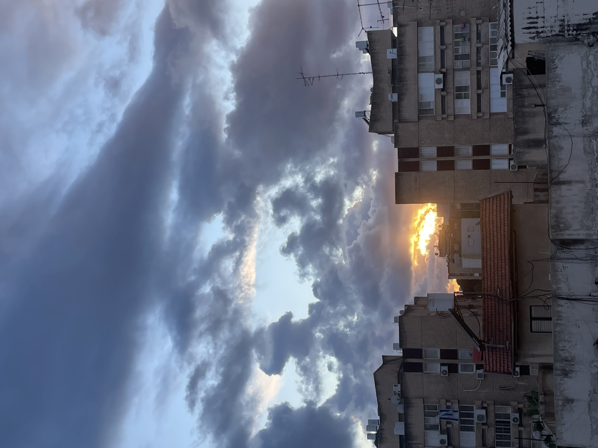The image displays an urban environment in Petah Tikva, Israel, at either sunrise or sunset, characterized by a dramatic sky and residential architecture. The upper two-thirds of the frame are filled with heavy, dark grey and purplish stratocumulus clouds, interspersed with clear areas of pale blue sky. A bright, golden-yellow light, consistent with the sun, is visible low on the horizon, partially obscured by clouds and structures.
In the midground, two multi-story residential buildings constructed of beige concrete flank a central space. The building on the left exhibits multiple windows equipped with white external Venetian blinds and numerous exterior air conditioning units. An Israeli flag, blue and white, is visibly suspended from an upper-floor window or balcony. Its rooftop features several white water tanks. The building on the right mirrors the construction style, also presenting windows with white blinds and air conditioning units. Its rooftop is equipped with a prominent multi-element antenna array, including a tall mast, alongside various white water tanks and satellite dishes.
A smaller, single-story structure with a red-tiled gabled roof is situated between the larger buildings in the lower midground. Mounted above this red roof is a solar water heater system, comprising a tank and a panel. The immediate foreground is defined by a weathered, grey concrete parapet or wall, with multiple electrical or utility wires attached. No human subjects, active interactions, or discernible motion are present in the scene.
