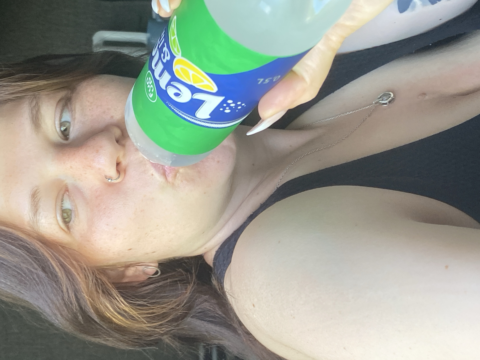 A woman is shown drinking from a bottle of Lemonade in Chaikovsky, Russia. The woman has long brown hair and freckles across her face. She is wearing a black tank top and a silver necklace. The bottle has a green and blue label with the text "Lemonade" and "0.5L" visible. The image appears to be taken indoors, possibly in a car, with natural light. The overall mood is casual and relaxed.