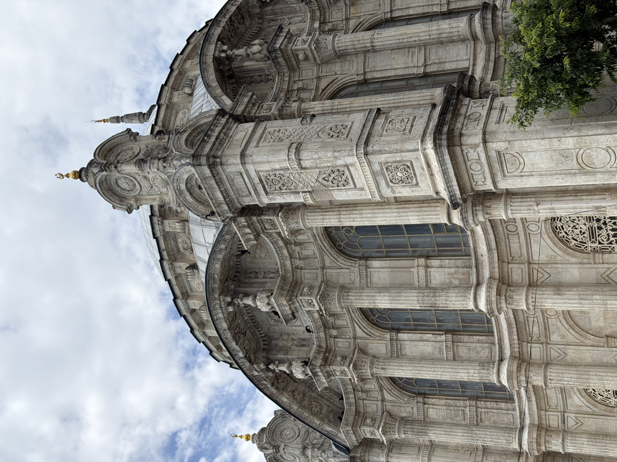 The image shows the exterior of the Ortaköy Mosque (Büyük Mecidiye Camii) in Beşiktaş, Turkey. It is a close-up, low-angle shot emphasizing the intricate architectural details of the mosque. The mosque is constructed of light-colored stone, featuring a combination of Baroque and Neoclassical design elements. There are visible columns, arches, ornate carvings, and window structures. The photograph captures the upper sections of the mosque, including the dome and a pointed spire. A small amount of greenery, likely from a tree, is visible on the right side. The background shows a cloudy sky.