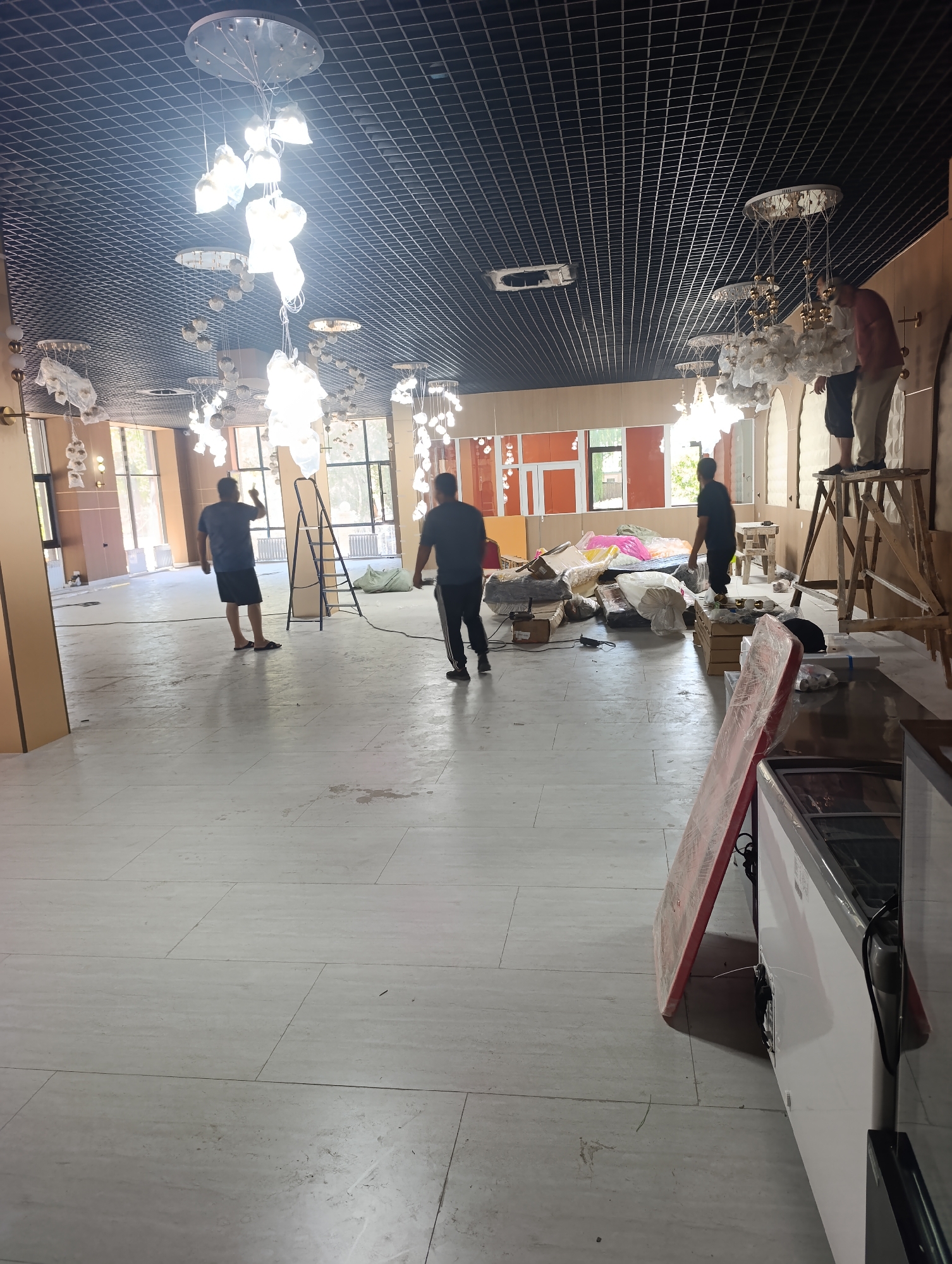 The image depicts the interior of a large, unfinished commercial space, likely a store or restaurant, with people engaged in setup or renovation work. The ceiling is covered with a black grid system and features numerous decorative pendant lights, some of which are partially wrapped. The floor is tiled with large, light-colored tiles, showing some dirt and scuff marks.
In the foreground, a red A-frame sign stands next to a white commercial refrigerator. The middle ground shows several individuals working. One person in a blue shirt and black shorts stands in the center of the room, gesturing with their right hand. To the right of this person, a stepladder is set up, and two individuals are visible on or near it, one wearing a red shirt and the other a dark shirt. Further to the right, another person in a dark shirt and black pants walks away from the camera.
The background reveals large windows looking out to greenery and daylight. The walls are painted in neutral tones, with some areas appearing unfinished. There are stacks of materials, including boxes, bags, and what appear to be furniture items wrapped in plastic, scattered throughout the space, indicating ongoing construction or unpacking. The overall impression is one of a business space in the process of being outfitted or renovated.