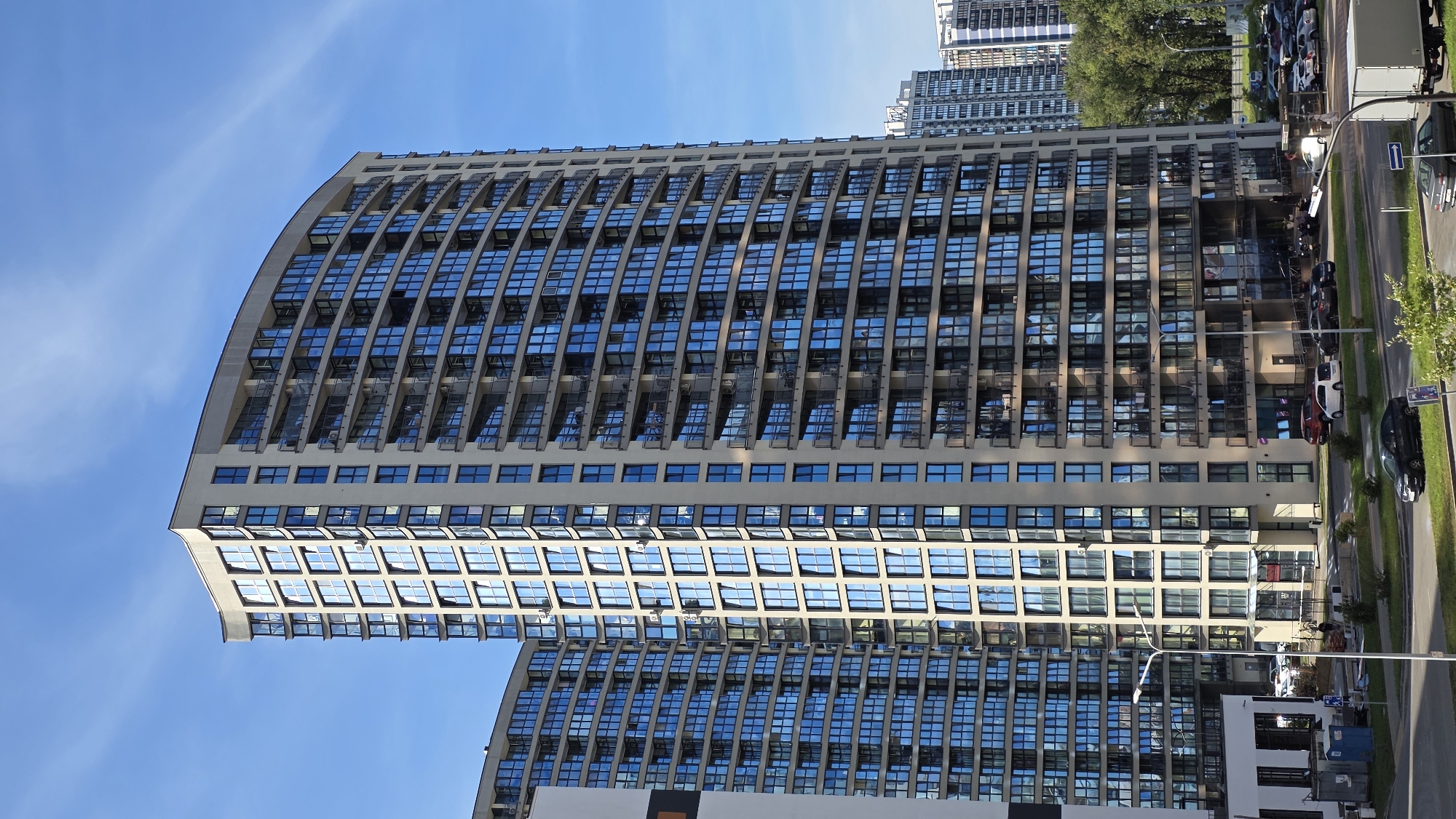 This is a photograph of a modern, curved apartment building with a beige facade and numerous reflective blue windows. The building has a slight convex curve and is several stories high, featuring balconies with dark railings on each level. To the left, another similar building is partially visible, also with many blue windows. The sky is a clear blue with faint wispy clouds. In the foreground, there is a street with several parked cars, including a white van, a red car, and a dark sedan. A lamppost stands near the sidewalk. Further in the background, other buildings of varying heights and architectural styles can be seen, along with some trees. The scene appears to be an urban residential area. The building in the foreground is the "Vivaldi House" residential complex, located in Minsk, Belarus, on Prospekt Pobediteley.