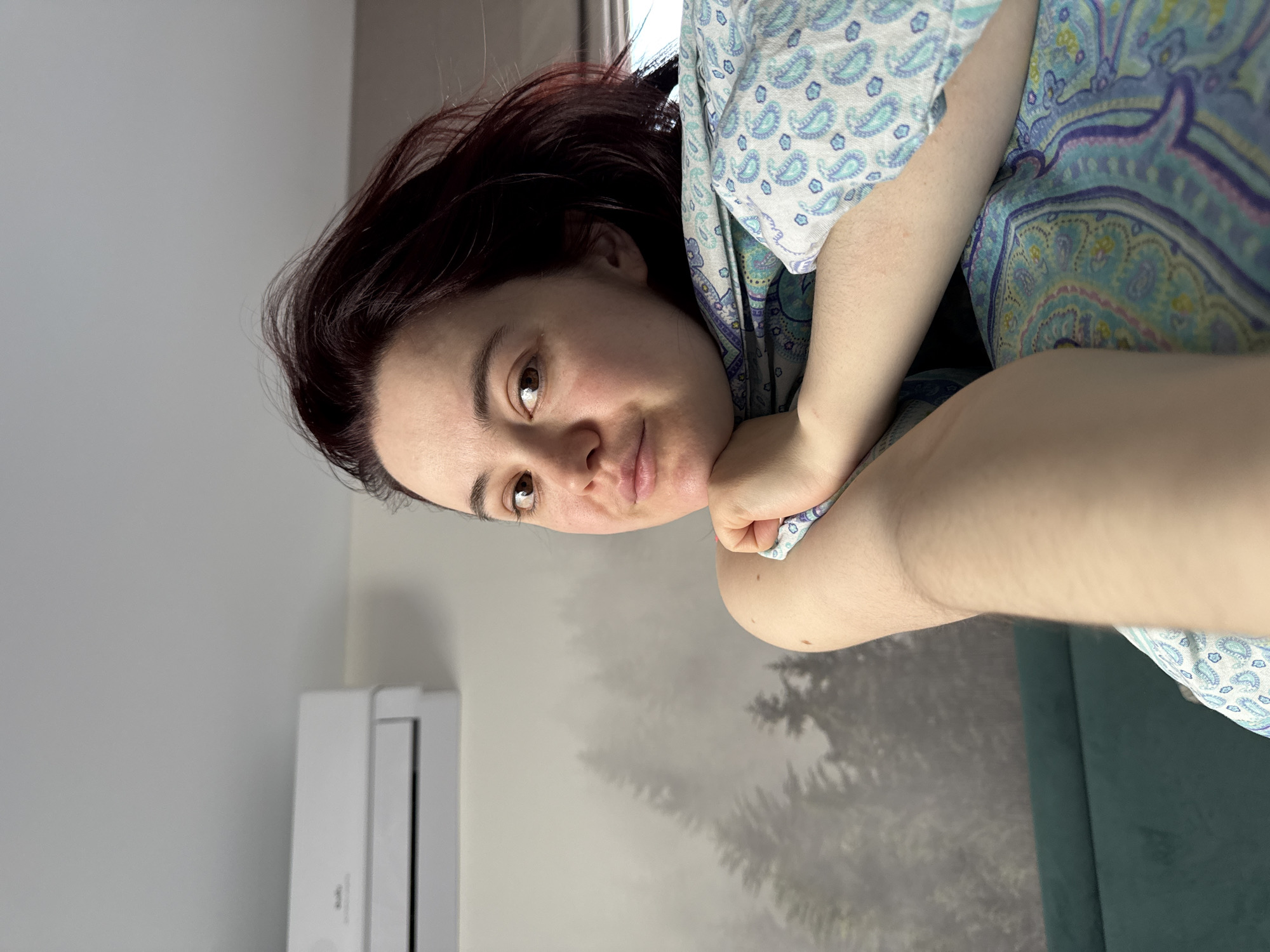 The image displays a woman captured indoors, with the photo oriented 90 degrees counter-clockwise from upright. The subject is a woman with dark, possibly reddish-brown hair and brown eyes, looking directly at the camera with a neutral to soft expression. Her head is tilted and appears to be resting on her right hand, supported by her left arm. She is dressed in a light-colored garment featuring a prominent blue and white paisley pattern.
The setting is a room, likely residential, in Izhevsk, Russia. The background consists of a light grey wall on the left, with a white, wall-mounted air conditioner unit partially visible in the lower left corner. Behind the woman, on the right side of the frame, is a decorative mural or wallpaper depicting dark, silhouetted trees in a misty or foggy forest scene against a lighter grey backdrop. A dark teal or green sofa or cushion is visible along the far right edge of the image. The lighting suggests it is daytime, providing soft illumination. No visible text is present in the image. The overall scene conveys a calm and relaxed atmosphere.