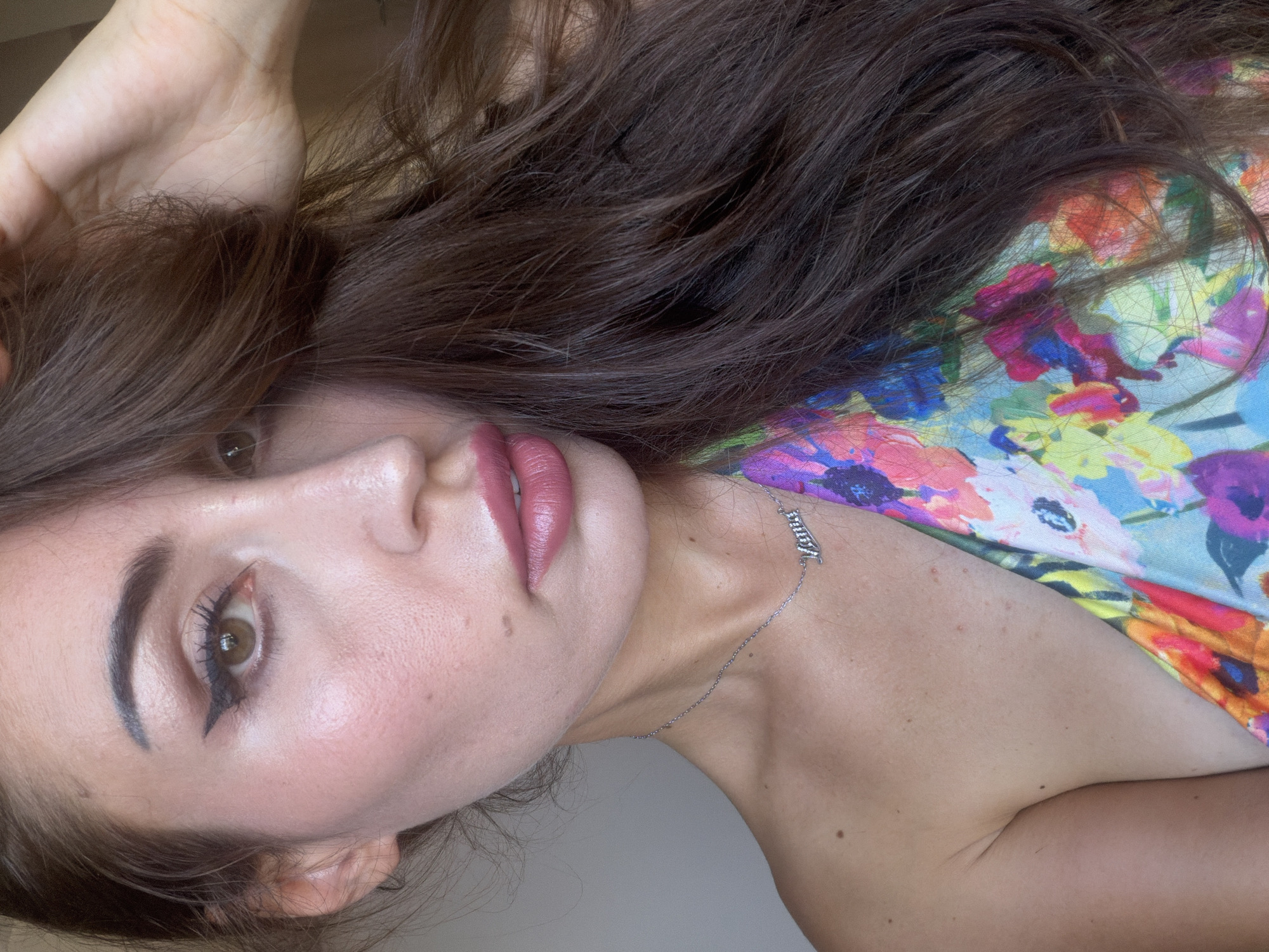A close-up, head-and-shoulders shot of a woman with long, dark brown hair is presented. She is wearing a brightly colored floral top and a silver necklace with the word "Lana" in script. Her makeup includes winged eyeliner and pink lipstick. Her left hand is touching her hair near her temple. The background is a plain, light-colored wall. The scene is set in Tersefanou, Cyprus.