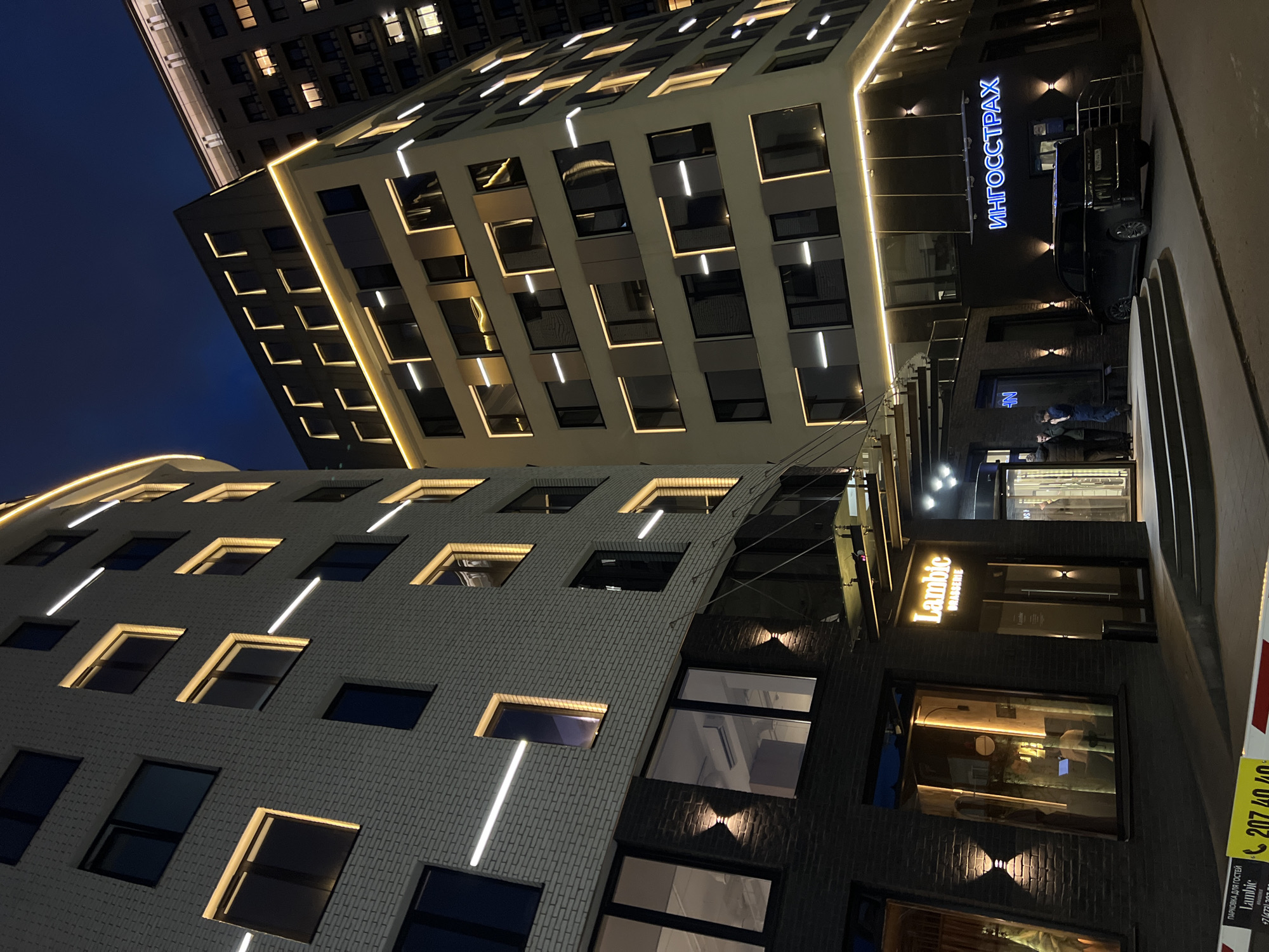 The image captures a vibrant urban night scene in Voronezh, Russia, showcasing modern architectural buildings illuminated against a dark blue sky. The main focus is on two multi-story structures, both featuring extensive exterior lighting.
The prominent building on the right has a light-colored facade, possibly textured, and is adorned with numerous linear LED light strips that outline its windows and architectural contours, creating a geometric pattern of light. A taller building is visible behind it, also lit from within and with some exterior lighting.
At street level, the base of the buildings hosts commercial establishments. A dark facade features a prominent blue neon sign spelling "ИНГОССТРАХ" (Ingosstrakh). A dark car is parked near this entrance. Further along the street, another illuminated sign clearly reads "Lambic BRASSERIE," indicating a restaurant or bar. Two people are visible standing near an entrance, possibly conversing.
Additional lighting includes uplights on the building facades and interior lights visible through the numerous windows. A yellow sign is partially visible in the bottom right, displaying "ПАРКОВКА ДЛЯ ГОСТЕЙ Lambic" (Parking for guests Lambic) and phone numbers, including "207 40 40" and "+7 (473) 201...", confirming the Voronezh location. The time of day is clearly night, with no indication of specific weather conditions other than a clear sky. The scene depicts a lively commercial area after dark.