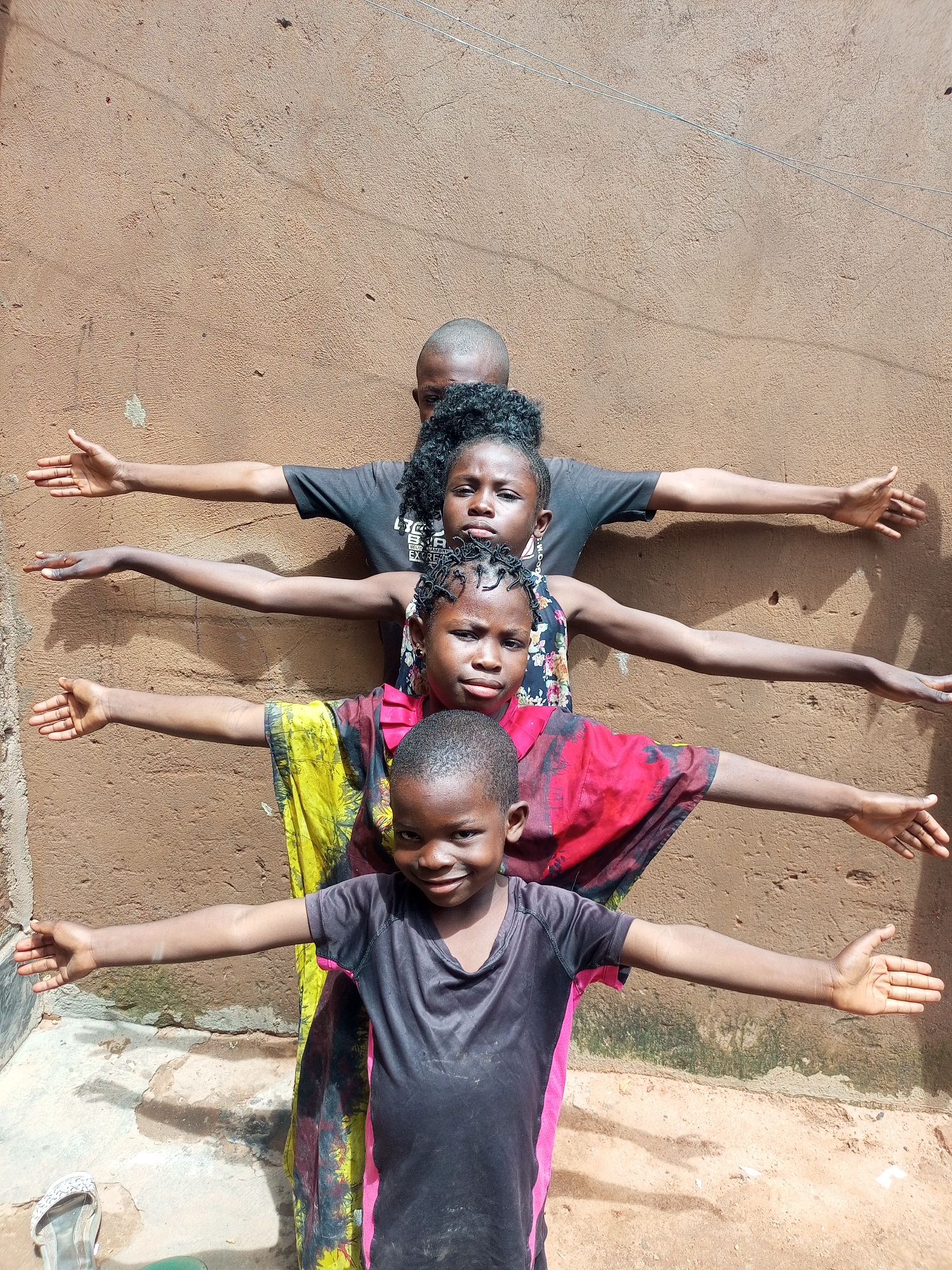 Four children stand in a line, facing forward, with their arms extended horizontally to their sides. The background is a textured, tan-colored wall. The child in the front, a young boy, wears a black t-shirt with pink trim. The child behind him, a girl, wears a red and black patterned garment over a yellow and green patterned fabric. The next child, a girl, wears a black t-shirt with a floral print. The child at the back, a young person with a shaved head, wears a black t-shirt. The ground in front of the children is a mix of dirt and concrete. There are faint shadows on the wall and ground.