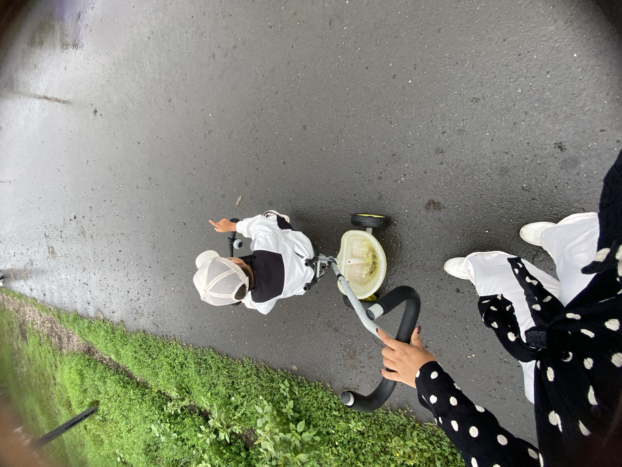 A child wearing a white cap and a black and white jacket rides a small tricycle on a wet asphalt path. The child is pointing with their right index finger. An adult's hands are visible on the handlebars of the tricycle, suggesting they are pushing or guiding it. The adult is wearing a black and white polka dot shirt and white pants. The scene appears to be outdoors, with a grassy area to the left of the path. The image is taken from a high-angle perspective, looking down on the child and the tricycle. The weather appears to be overcast or rainy, as the asphalt is wet and reflective. The specific location in Maloarslangulovo, Russia, is not visually evident from the image itself but is provided as context.