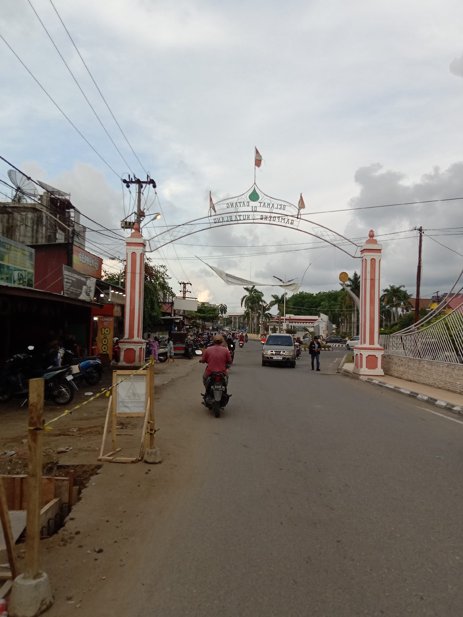 The image depicts a street scene in Lhokseumawe, Indonesia. A road runs through a red-and-white entrance archway that reads "SELAMAT DATANG DI KOTA LHOKSEUMAWE" in mirrored letters. The sky is overcast. Various motorbikes and a silver van are on the road, with a man walking near the van. Shops and buildings line the left side of the road. There is construction on the left edge of the image, indicated by wood barriers and yellow caution tape. Power lines are visible overhead. Palm trees and other greenery are in the background.