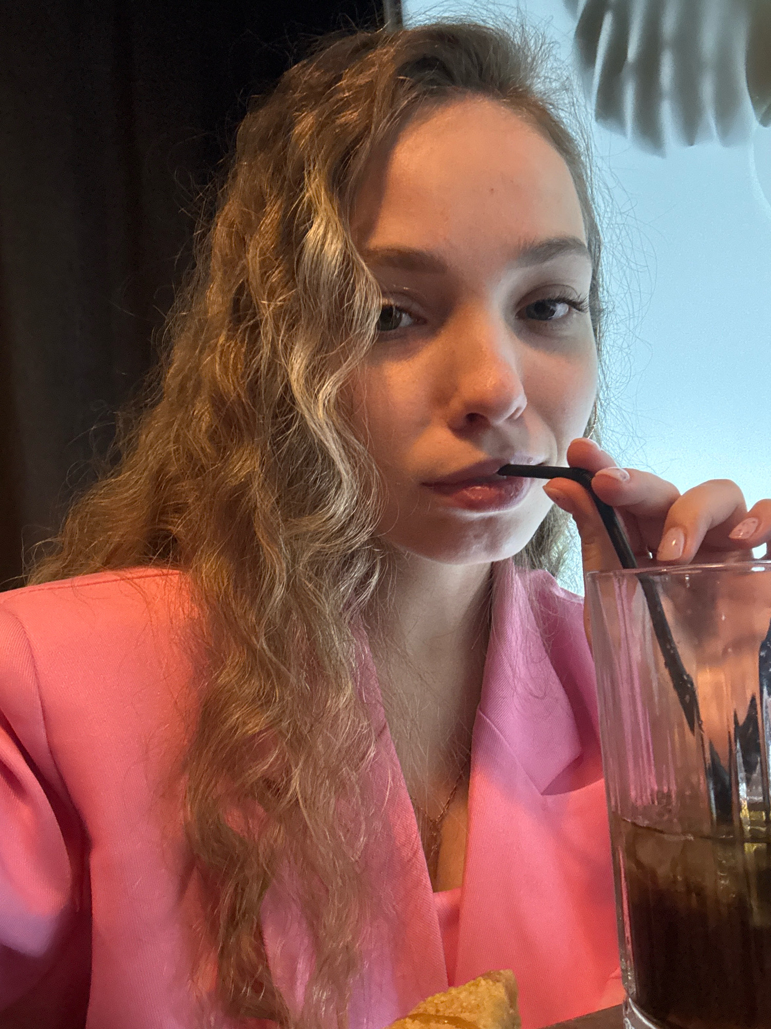 In a dimly lit indoor setting, a young woman with long, wavy blonde hair is captured in a close-up, selfie-style shot. She is wearing a vibrant pink blazer over a darker top, and a delicate necklace is visible at her throat. Her left hand, with manicured nails, holds a black straw to her lips, suggesting she is drinking from a tall glass filled with a dark beverage and ice. The background is divided between a dark, textured area on the left and a brightly lit, possibly window or screen, on the right, which features an abstract, white, wavy pattern. The overall ambiance suggests a casual dining or social setting, potentially a restaurant or cafe. The location of Chaikovsky, Russia, is not visually discernible from the provided image.